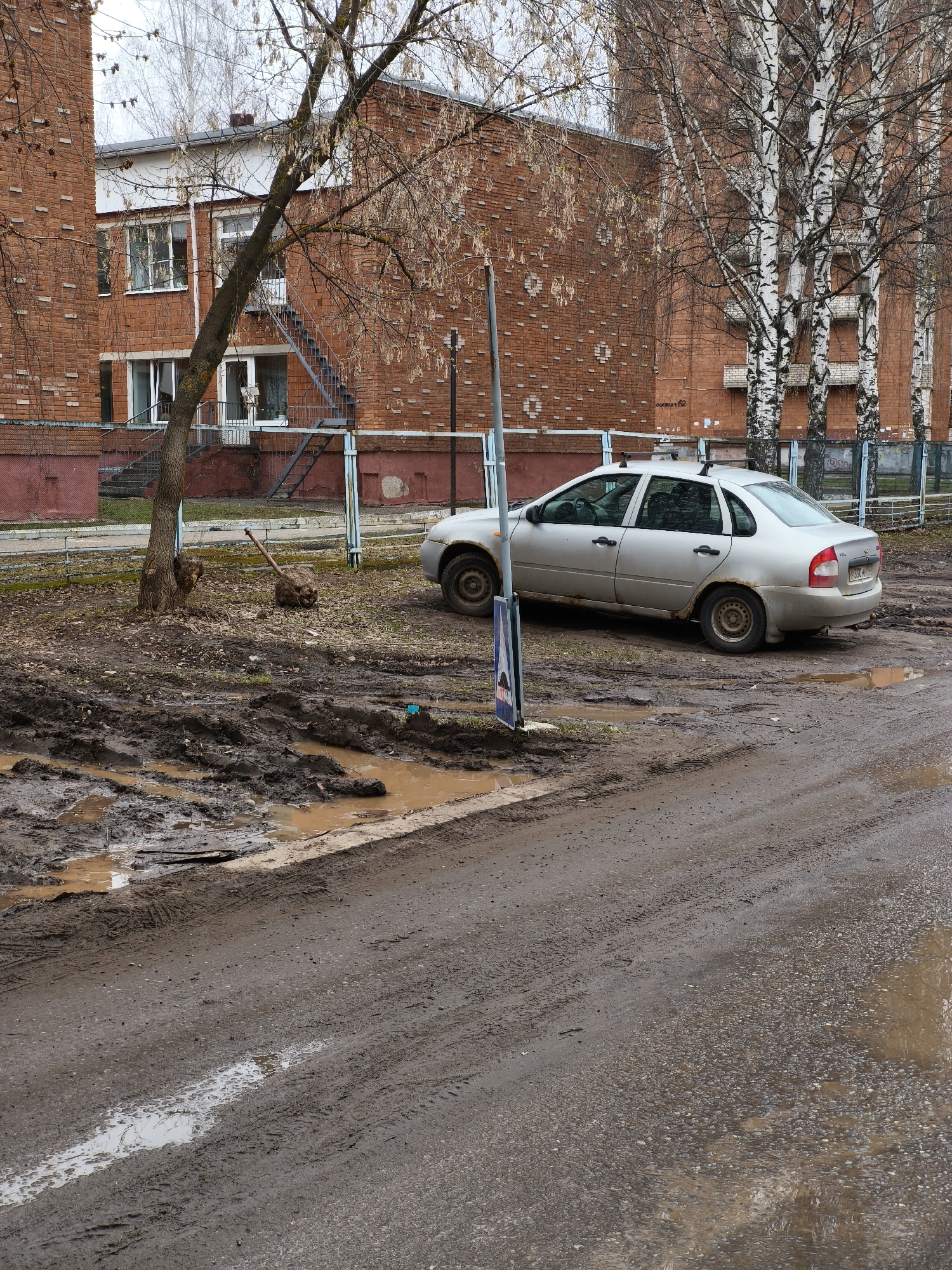 The scene is a muddy street in Izhevsk, Russia, with a silver Lada sedan parked adjacent to a sidewalk and next to a tree. A red brick building is behind the car. The ground is covered in mud and puddles. A street sign is adjacent to the car's front left. A tool (axe) is leaned against the tree. Bare birch trees are next to the red brick building. The weather is overcast.