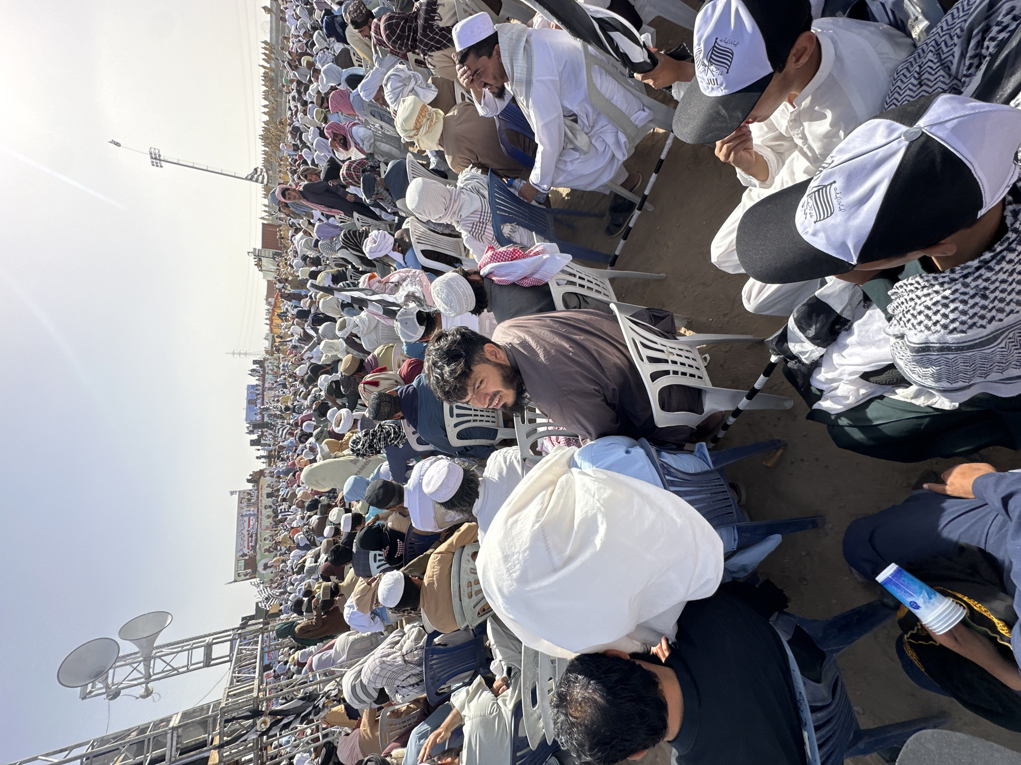 The image depicts a massive outdoor gathering of men, possibly hundreds or thousands, seated on plastic chairs across a large, dry, and dusty field in Pishin, Pakistan. The scene is illuminated by bright, clear daylight, suggesting a daytime event.
The primary subjects are men of varying ages, predominantly dressed in traditional South Asian attire such as shalwar kameez. Many wear kufis (prayer caps) or turbans in white, beige, or checkered patterns, reflecting the conservative religious and cultural context. Several individuals, particularly prominent in the foreground and visible in crops, wear black and white baseball caps bearing the flag symbol and Urdu text "جمعیت علماء اسلام" (Jamiat Ulema-e-Islam – JUI), a major religious-political party in Pakistan. A large banner or stage in the distance also displays text and portraits consistent with JUI, indicating the event is likely a political rally or religious congregation organized by the party.
Attendees are mostly facing forward, suggesting they are listening to speeches or participating in a collective activity. While many appear attentive, some are seen resting their heads, shielding their eyes from the sun, or looking tired, reflecting the long duration or warm weather conditions. One man in the foreground wears a brown shirt and has a slight smile, looking towards the camera.
In terms of objects, numerous white and blue plastic chairs fill the frame. Large speaker systems are mounted on scaffolding, designed to project sound over the extensive crowd. A blue plastic cup is visible on the ground near one attendee. The overall setting is functional and geared towards accommodating a very large audience in an open-air environment.