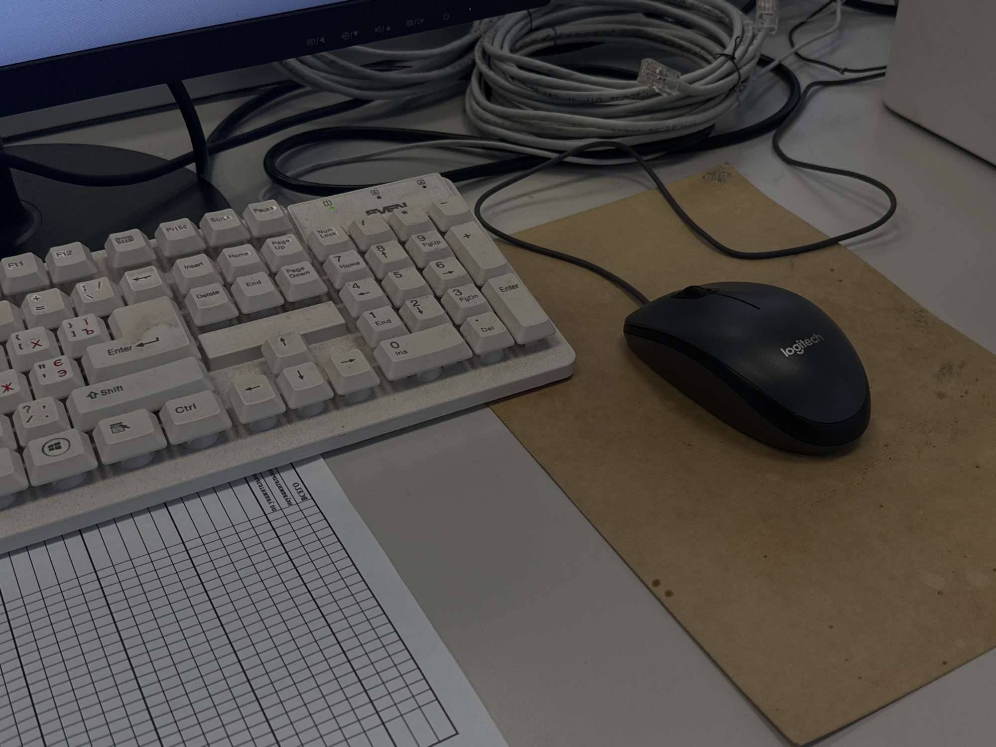 The image depicts a workspace with a computer monitor at the top, partially visible. Below the monitor, several coiled grey network cables are arranged. A white keyboard, labeled "SVEN," is positioned centrally. A black Logitech mouse rests on a brown, worn surface, possibly a mousepad. To the left of the keyboard, a sheet of paper with a grid pattern and text in what appears to be Russian is visible.
Based on the presence of Russian text on the document and the keyboard layout, the location is likely within Russia. Without additional information, it's not possible to confirm if this image was taken in Perm, Russia.