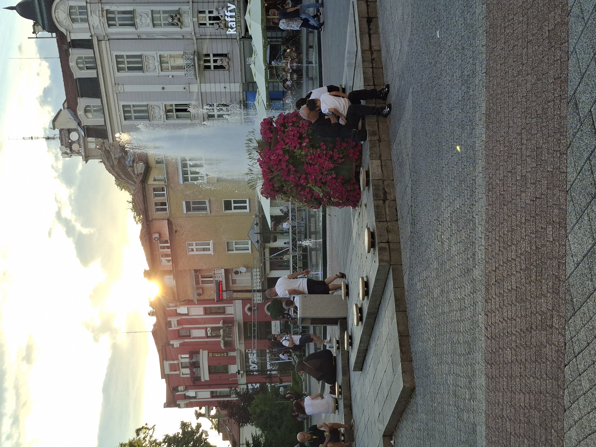 The image shows a scene in the city of Plovdiv, Bulgaria, specifically featuring a fountain and surrounding buildings. There are several people present in the vicinity of the fountain; some are seated on its edge, while others are walking around. One group consists of an adult and child near a large flower planter, while others are near the water. The environment includes buildings with architectural details typical of European cityscapes. A restaurant called "Raffy" is identifiable in the background. The sky is bright, with the sun appearing to be setting. The ground is paved with stones.