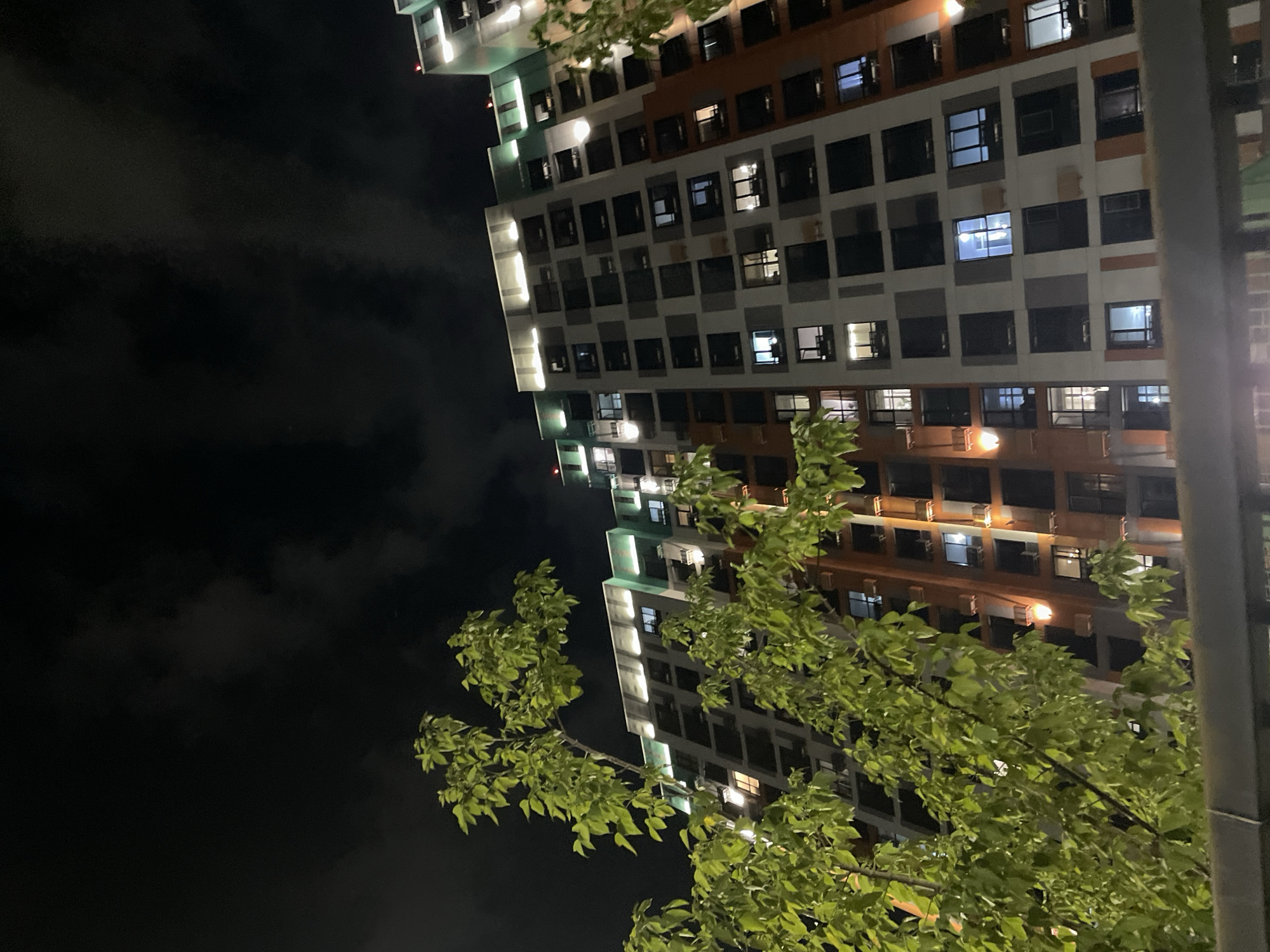 The image shows a modern high-rise apartment building at night, viewed from a low angle through the branches of a tree with bright green leaves. The building is illuminated by internal lights visible through its numerous windows, and by exterior lighting along its upper levels. The sky is dark with clouds. The building features a multi-colored facade, with sections of white, gray, and terracotta. The trees in the foreground partially obscure the view of the building, creating a sense of depth and framing the scene. The overall impression is one of urban residential life at night, with the natural element of the tree providing a contrast to the man-made structure.