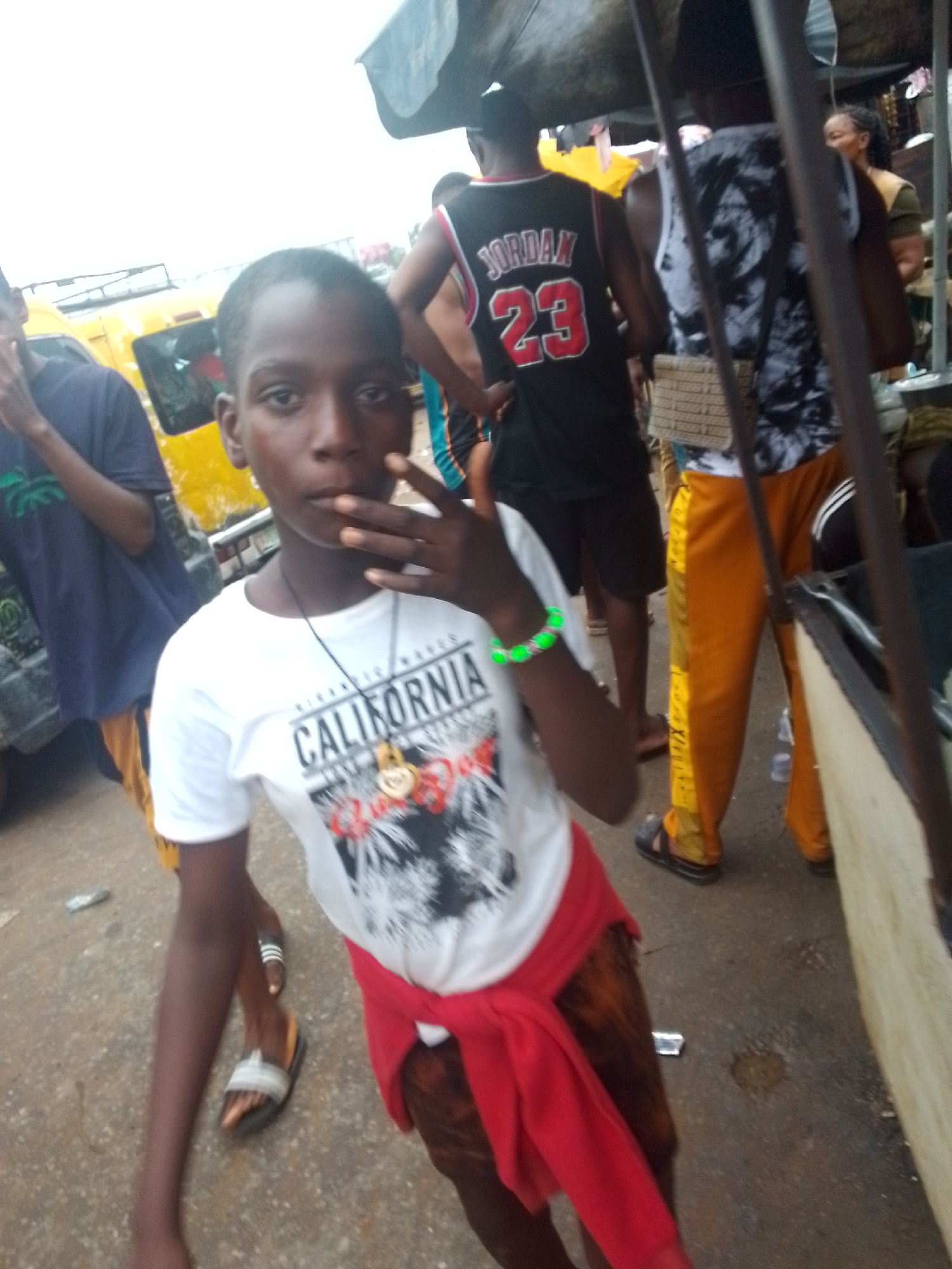 In Nkpor, Nigeria, a young Black individual is prominently featured in the foreground, gazing directly at the camera with a hand near their mouth. They are wearing a white t-shirt with the word "CALIFORNIA" printed on it, a red cloth tied around their waist, and dark brown pants. The individual has a necklace with a gold pendant. To the left, another individual in a dark blue t-shirt and brown shorts is partially visible. In the background, a yellow vehicle, likely a bus, is parked. Further back, two other individuals are present: one wearing a black "JORDAN 23" basketball jersey and another in a patterned tank top and orange pants. The scene appears to be outdoors, possibly a street market or roadside setting, with overhead structures and other people in the distance. The ground is paved.