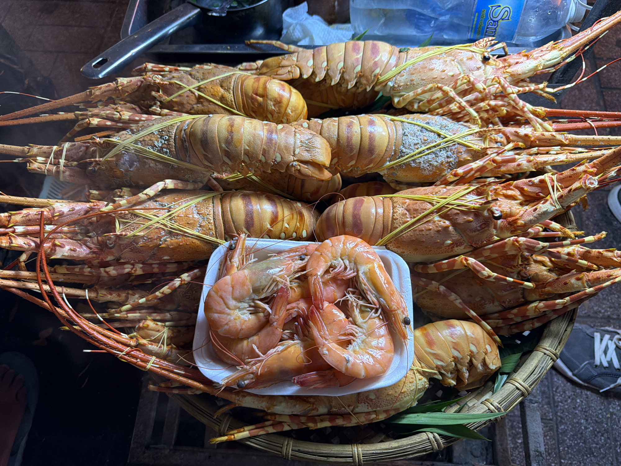 The image shows a street food display in Nha Trang Ward, Vietnam. A woven basket is filled with cooked spiny lobsters, identified by their banded legs and distinctive body segments. They are arranged in layers, some bound with thin, yellow string. A white styrofoam container filled with cooked, orange-pink shrimp sits atop the lobsters. Steaming suggests the seafood is freshly cooked. A stainless steel serving utensil is visible at the top left. Part of a "San" branded water bottle is visible in the background. The edge of a person's sandal-clad foot and a sneaker-clad foot are visible on either side of the basket, suggesting a vendor and potential customer in close proximity. The street surface is visible underneath the basket.