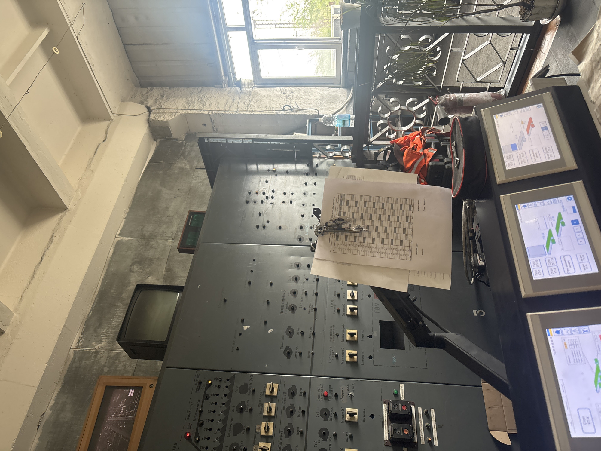 This media file captures a detailed view of a control panel within an industrial or technical setting. The panel is primarily composed of a large, dark gray console covered in numerous buttons, switches, and indicators, suggesting it's part of a complex operational system. Several labels in Cyrillic text are visible on the panel, including terms like "ДИСПЕТЧЕР" (dispatcher), "АСЛЕТЧРЕВ" (likely related to lubrication or oil systems), and station designations like "СТАНЦИЯ ПРИВОДОВ З" (Station of Drives 3).
Two modern touch-screen monitors are prominently displayed at the top right of the console, showing graphical interfaces with data and controls, further indicating a technologically advanced system. One screen displays a diagram with numerical values and operational states, while another shows a map-like interface with graphical elements.
To the left of the main console, an older, smaller CRT monitor is visible, adding a sense of historical depth or a hybrid operational setup. In the background, there is a large window offering a glimpse of an outdoor scene with greenery, suggesting daylight and a possible connection to the outside environment. The overall impression is one of a functional, possibly operational, control room. The presence of safety vests and other equipment suggests an active work environment. The context of Kryvyi Rih, Ukraine, implies this could be a control center for mining, industrial processing, or infrastructure operations in that region.