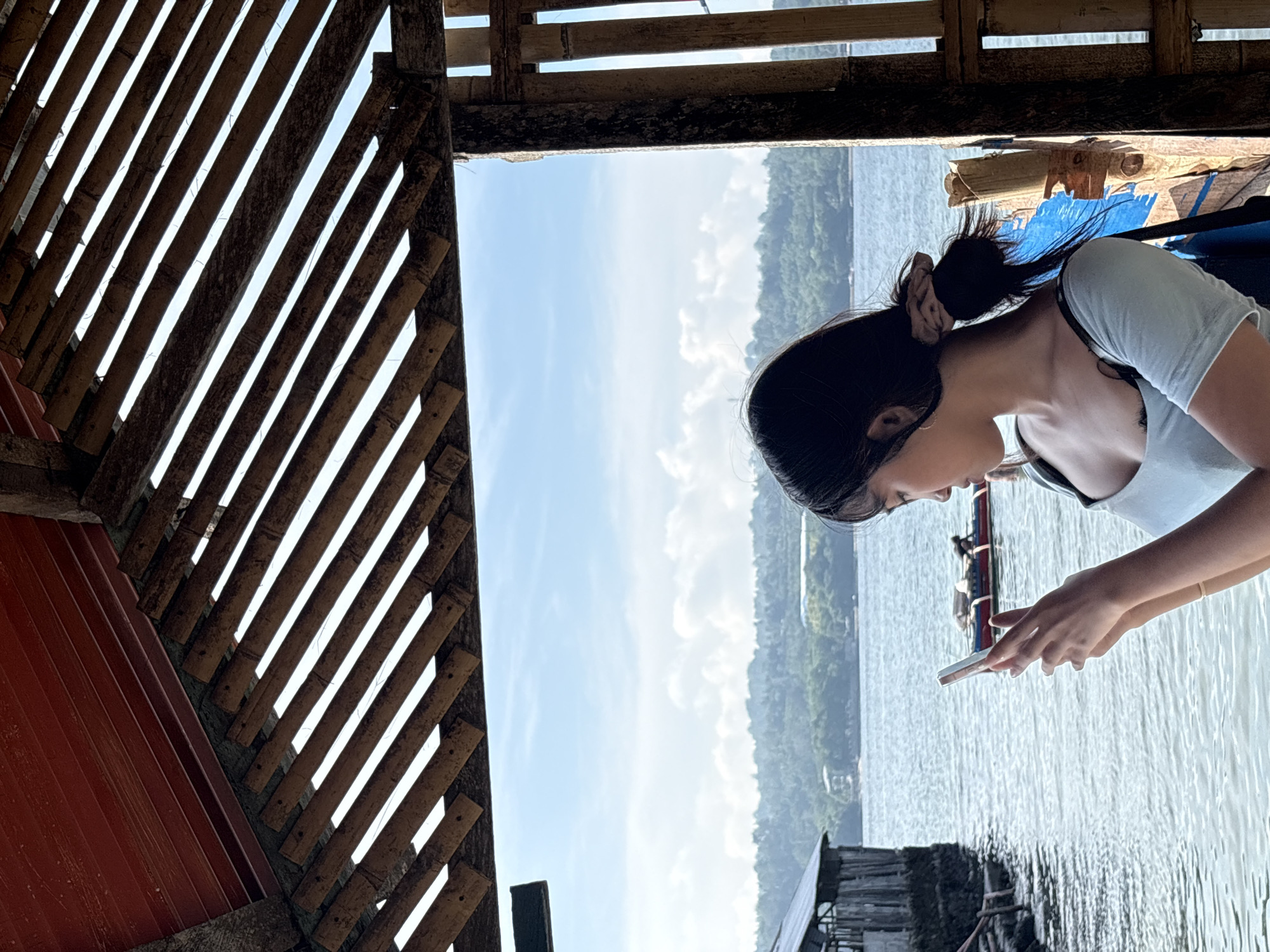 A young woman with dark hair tied back in a bun is looking at her phone. She is wearing a light blue top with a black strap visible. She is standing on a wooden platform, possibly a pier or a hut, with a thatched roof made of bamboo. In the background, there is a body of water, likely the sea, with a small boat in the distance. The landscape beyond the water appears to be hilly and covered with vegetation, suggesting a tropical environment. The sky is blue with scattered clouds. The presence of a boat and the lush green hills suggest a coastal area. This scene is consistent with many picturesque locations found in Davao City, Philippines, particularly along its coastline which offers similar vistas of the sea and surrounding islands or mainland.
