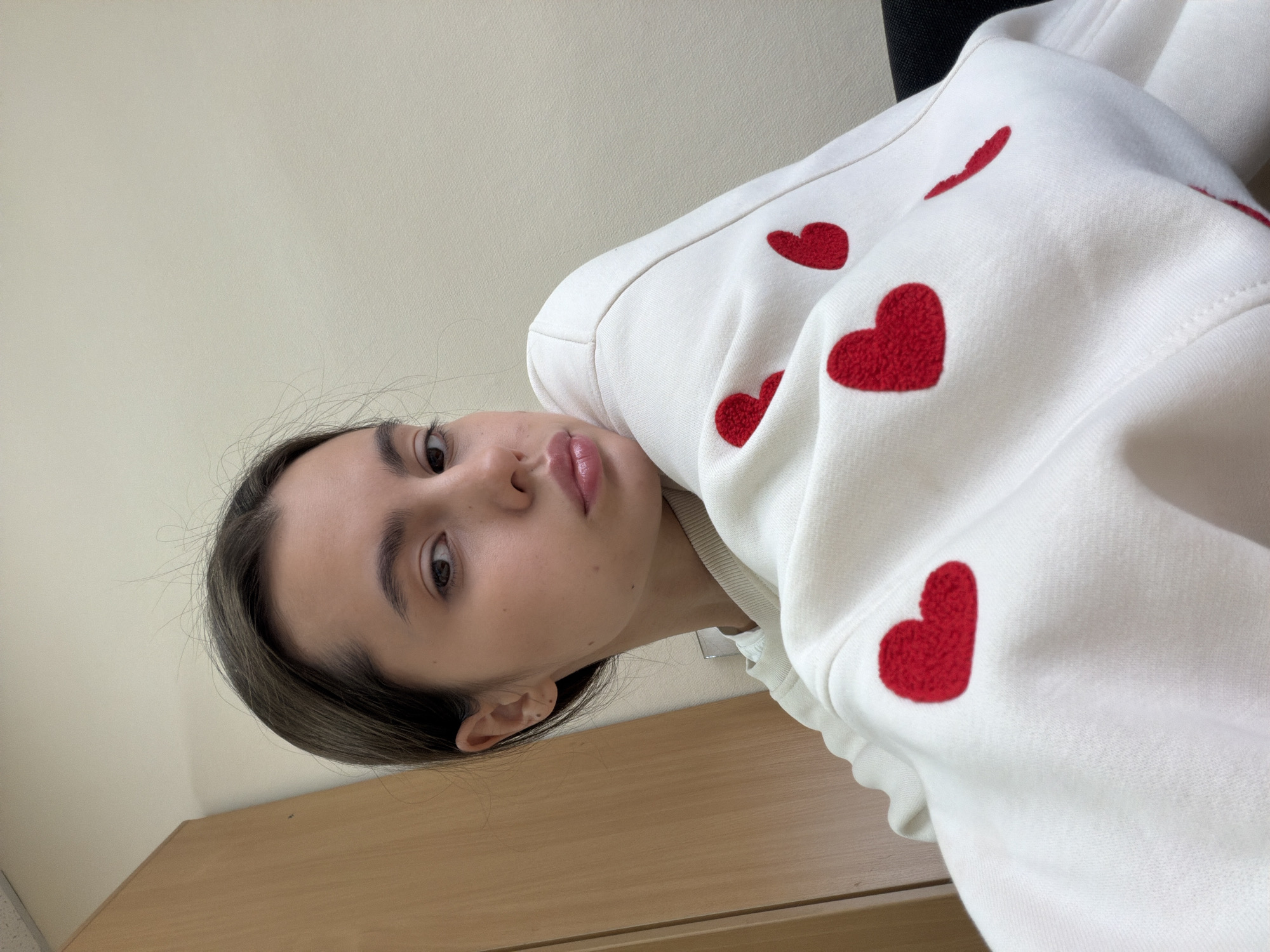 The image captures a selfie of a young woman, rotated 90 degrees clockwise, suggesting she is either lying down or tilting her head significantly. She has dark hair, a neutral expression, and is looking directly at the camera. Her lips are slightly parted. She is wearing a white long-sleeved garment, likely a sweatshirt or hoodie, which is adorned with multiple textured red heart-shaped appliques or embroidery.
The setting appears to be indoors. A plain, light-colored (possibly beige or off-white) wall forms the background in the upper portion of the frame. A wooden surface, resembling a desk or table, is visible in the lower part of the image. A dark, indistinct object, possibly a chair, can be seen behind her right shoulder. There is no visible text in the image. The image provides no specific cues for the time of day or weather, nor for the location in Chaikovsky, Russia beyond the given context.