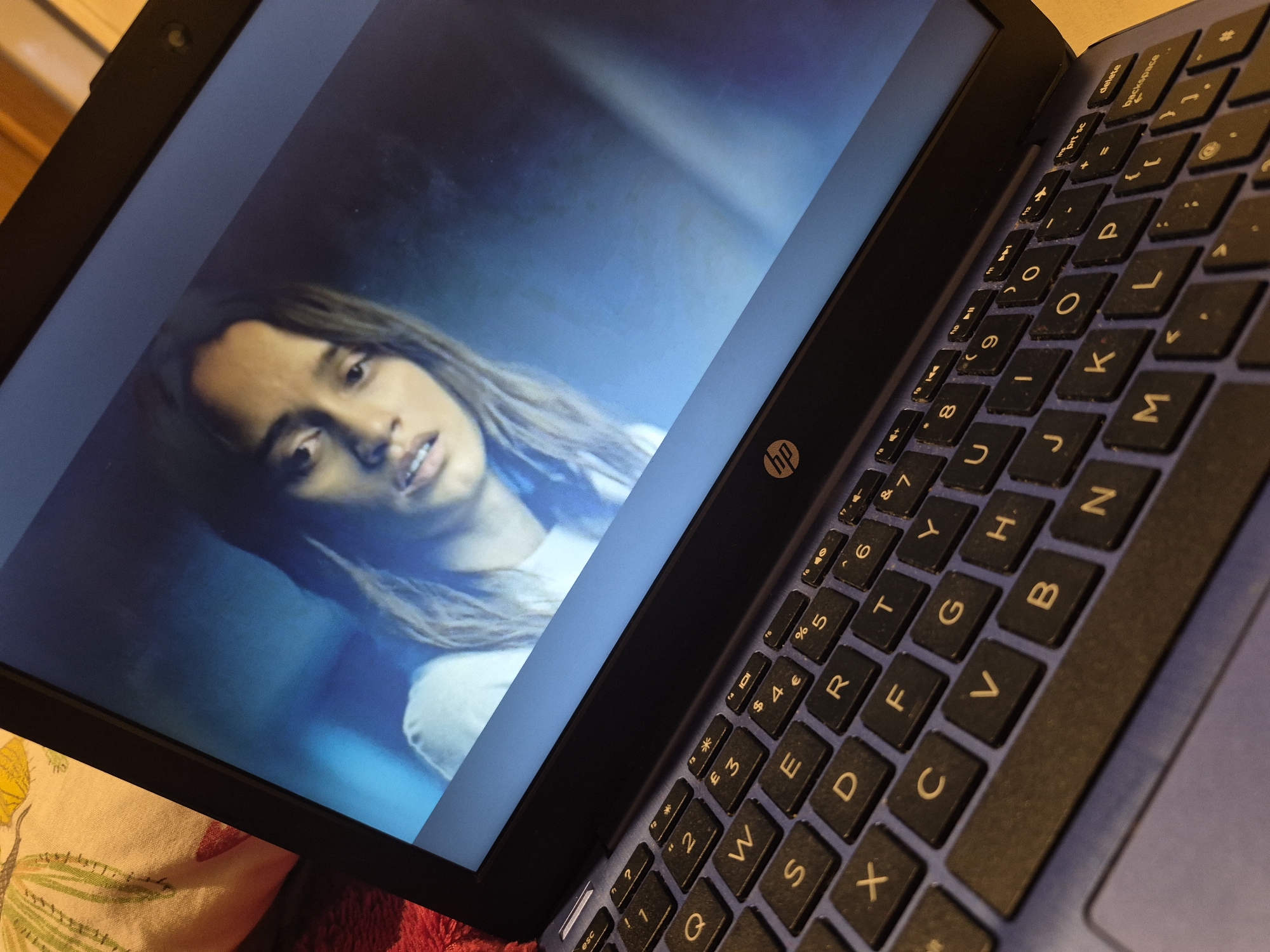 An HP laptop is positioned on a red textured surface, angled to display its screen. The screen shows a close-up of a young woman with brown hair, looking distressed with her mouth slightly open. The background on the screen is a dark, blurred blue. The laptop's keyboard is visible and is blue, with black keys featuring white lettering. Some keys are identifiable, including "esc", "?", "1", "2", "€", "3", "W", "S", "D", "E", "R", "4", "%", "5", "6", "&", "7", "X", "C", "F", "T", "Y", "8", "U", "I", "O", "P", "{", "}", "delete", "backspace", "B", "N", "M", "L", and "K". The HP logo is visible on the laptop's bezel. In the upper left corner of the image, part of a patterned cloth with yellow and green floral designs is visible, resting on a beige surface. The overall lighting suggests an indoor setting, potentially evening or low-light conditions, with the laptop screen being the primary light source. There is no information in the image to suggest a location in the city of Sandwell, United Kingdom.