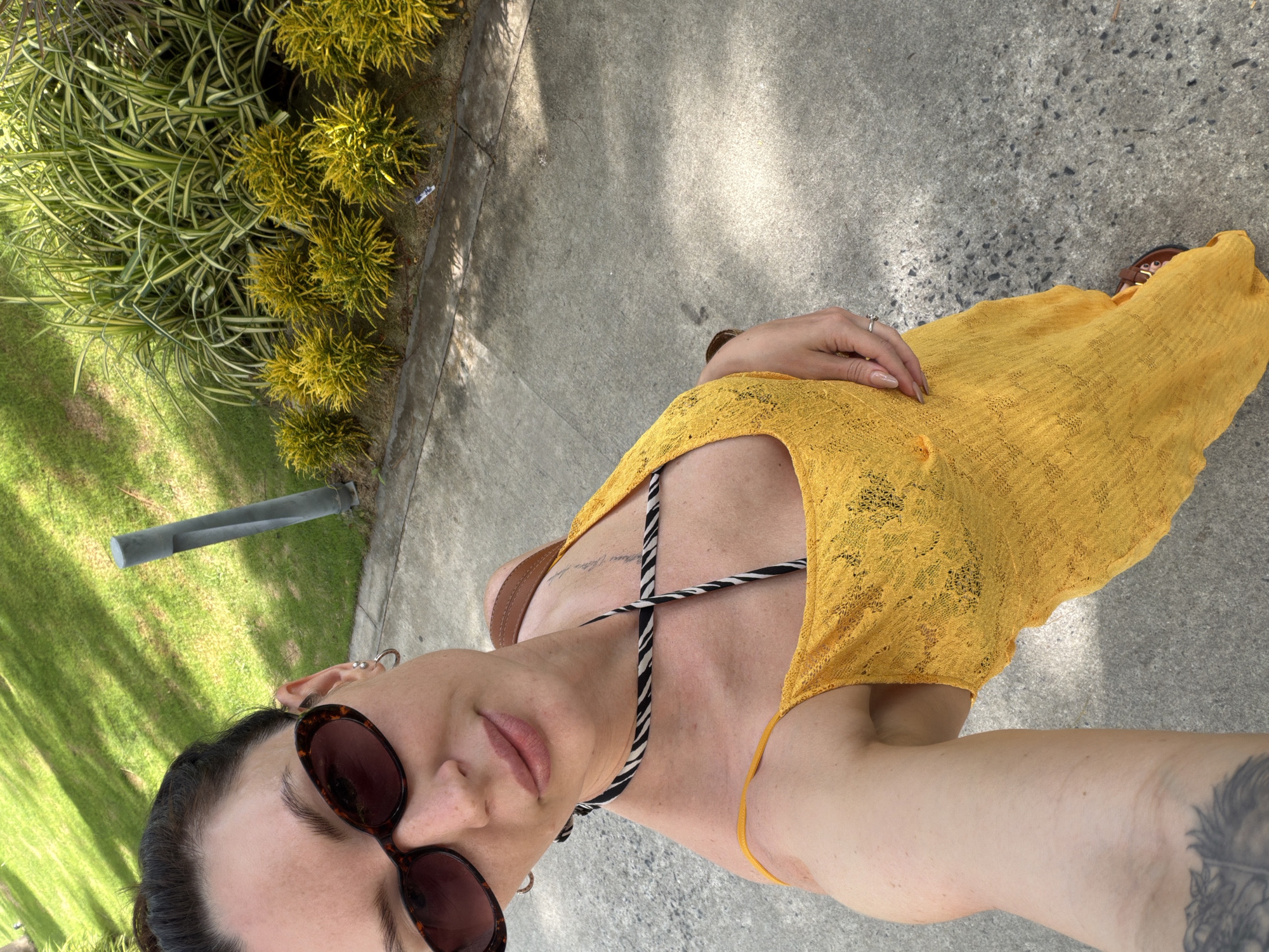 A woman is taking a selfie outdoors. She is wearing a yellow dress, sunglasses, and a black and white striped necklace. The background shows a lawn, a path, and some bushes. The location is Dong Ninh Hoa Ward, Vietnam.