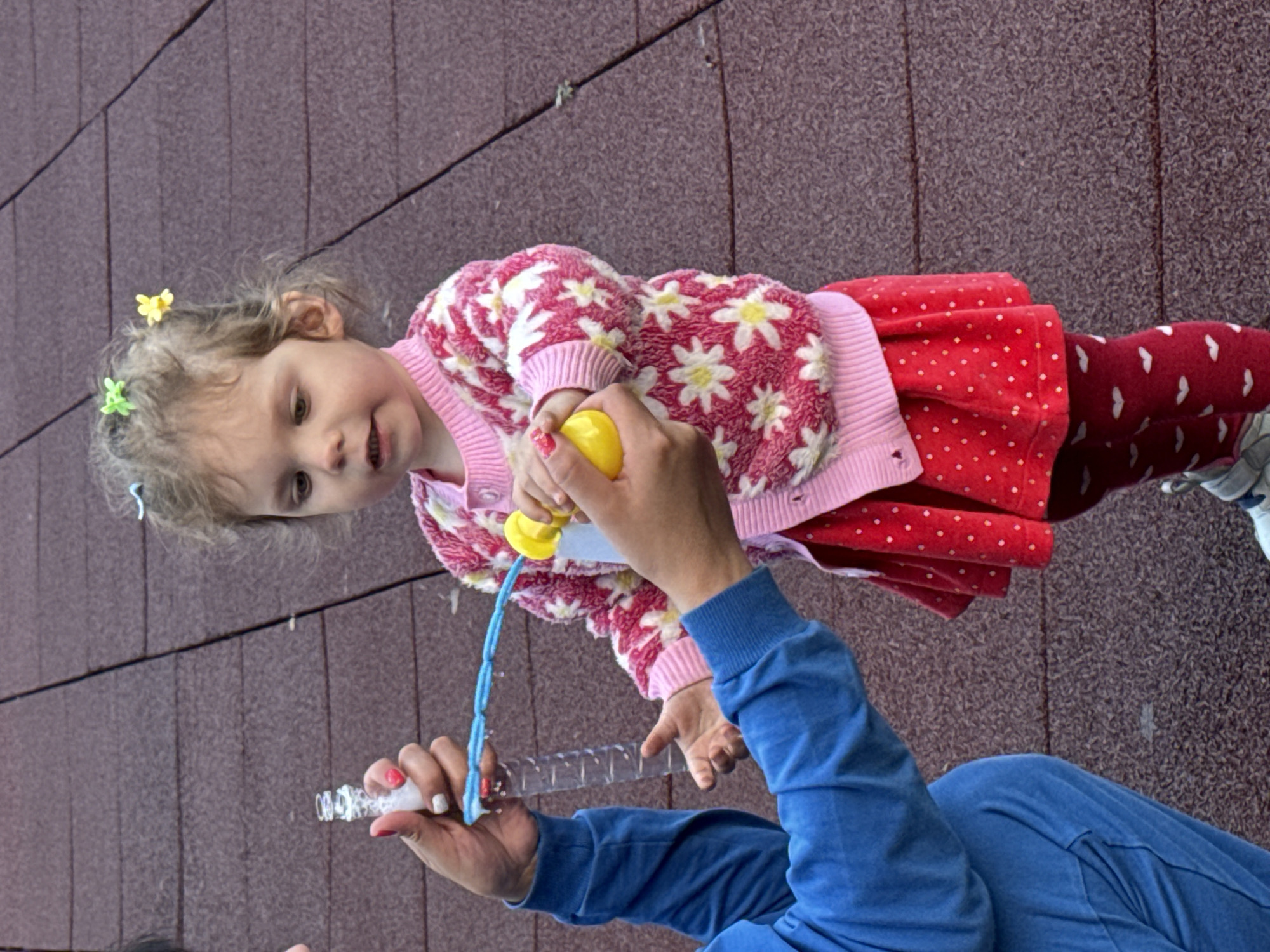 A toddler with curly blonde hair, adorned with colorful flower-shaped hair clips, stands on a reddish-brown tiled surface. She is wearing a pink cardigan with a floral pattern and a red polka-dot skirt. Her legs are covered in red leggings with white heart patterns. To her left, an adult, wearing a blue tracksuit, is holding a plastic bottle of bubble solution and a blue bubble wand. The adult's hands are near the child's face, and the child is looking forward with her mouth slightly open, appearing to be about to blow bubbles. The background is a textured, reddish-brown surface that appears to be part of an outdoor play area.