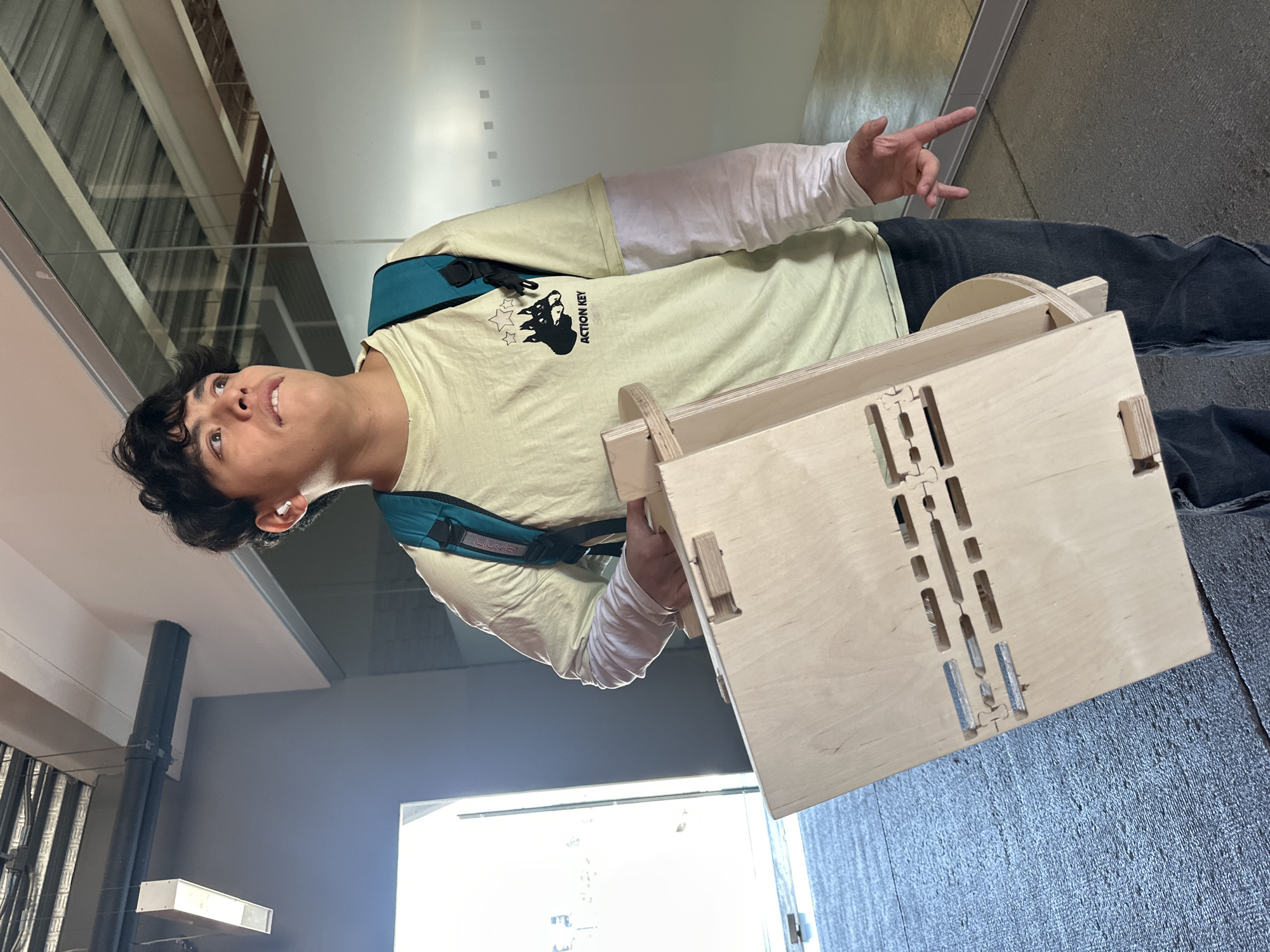 A young man stands indoors, holding a light-colored wooden box with rectangular cutouts on the side. He wears a pale yellow t-shirt with a black logo and the words "ACTION KEY" printed on it, a white long-sleeved shirt underneath, and dark pants. A turquoise backpack strap is visible over his shoulder. He is looking up and to the left, gesturing with his left hand.
The environment appears to be a modern, industrial-style building, with exposed ceilings and piping, and a view to an outdoor area with greenery. The location is likely in Mexico City, Mexico.