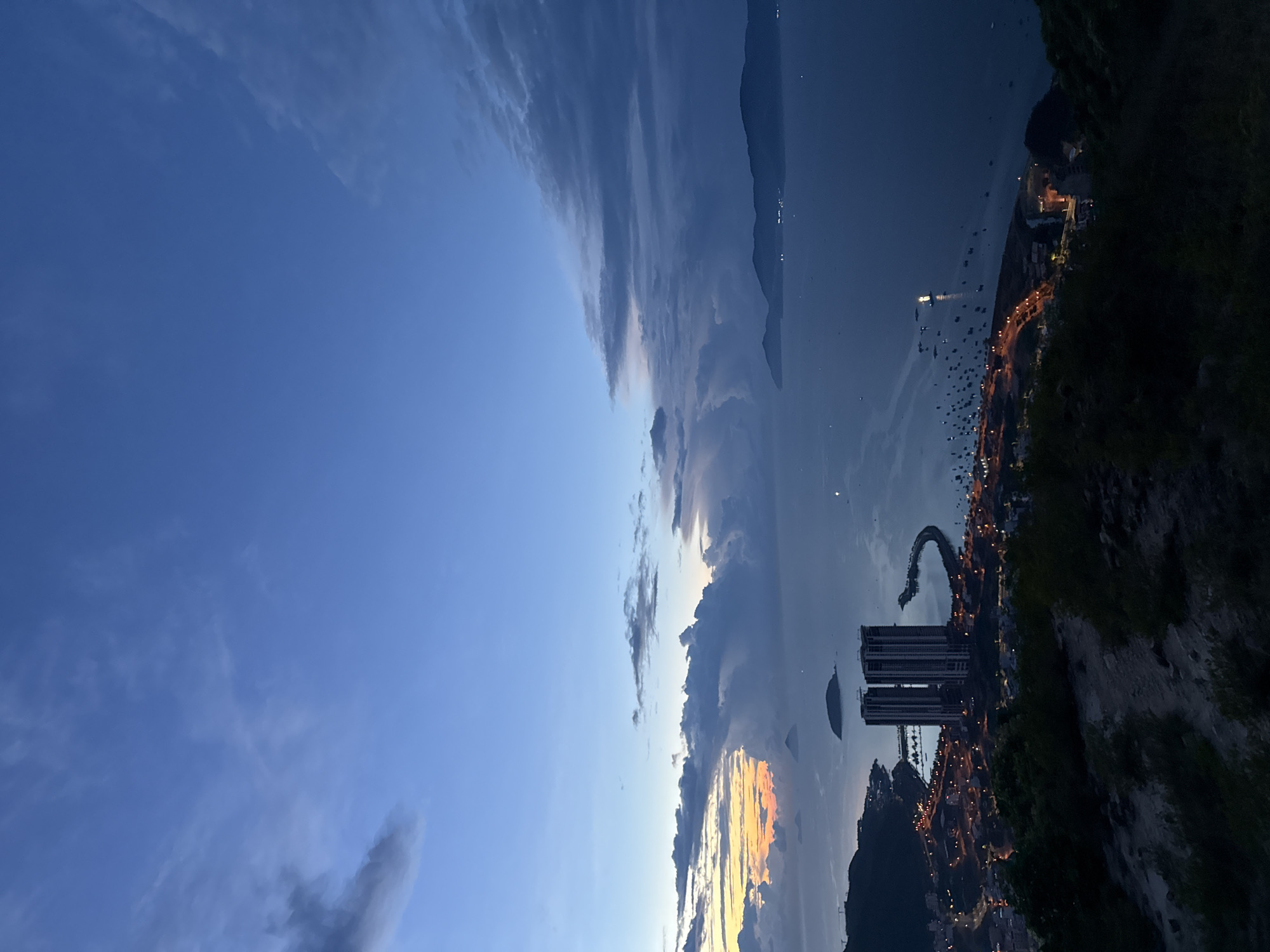 The image depicts a twilight scene in or near Bac Nha Trang Ward, Vietnam, with the sky occupying the upper portion of the frame. The sky transitions from a clear, deep blue to a mix of clouds tinged with sunset colors.
Below the sky, a city or town is visible along the coastline. Lights from buildings and streetlights illuminate the area, suggesting a residential or urban setting. Several boats are docked in the water.
The lower right corner of the image shows a steep, dark hill or mountain covered in vegetation. Modern architecture is visible in the city.
The image is presented sideways.