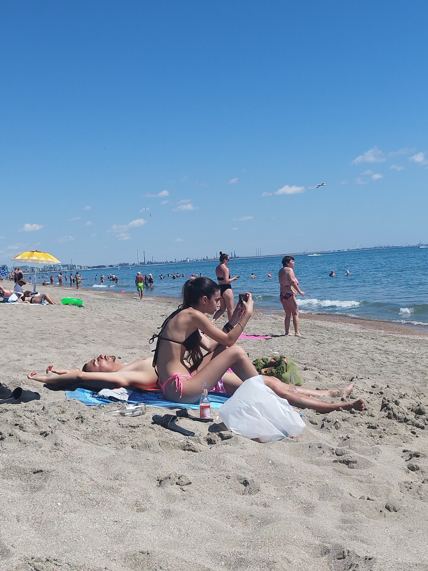 The image depicts a sunny day at a crowded beach, likely in Constanța, Romania, given the industrial skyline visible in the distance. In the foreground, a man lies on his back on a towel, arms outstretched. Beside him, a woman sits with her back to the camera, holding a phone and appearing to take a picture. Both are wearing swimwear. Further back, other beachgoers are visible, some in the water, others on the sand, and some under a yellow umbrella. The sea is calm with small waves, and the sky is a clear blue with a few scattered clouds. There is also a faint impression of an airplane in the sky. The overall scene captures a typical summer day at the seaside.