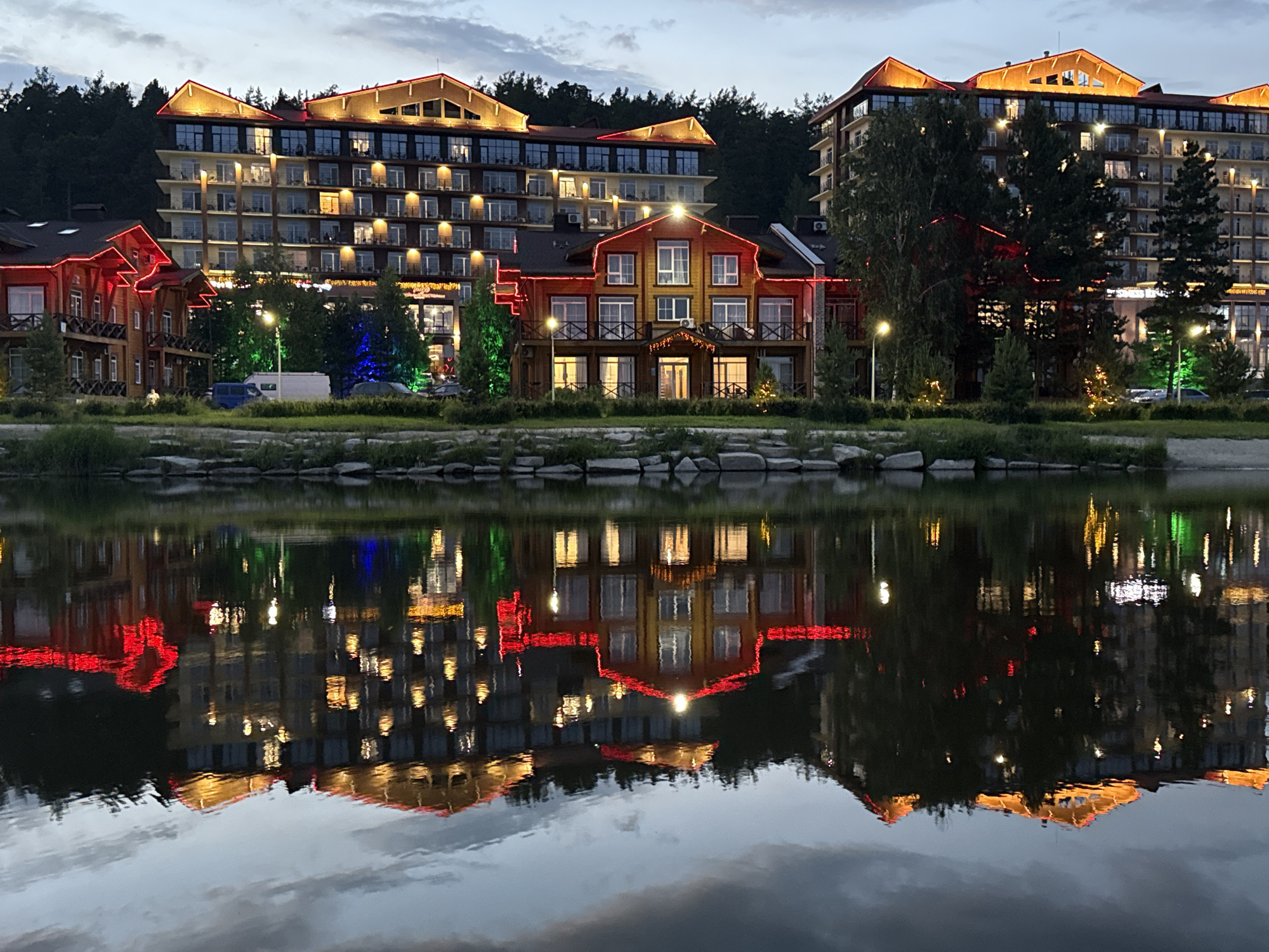This is a nighttime, wide-angle, full shot of the Syrostan Resort in Syrostan, Russia, featuring several multi-story buildings and their reflections in a body of water. The buildings are lit with warm, yellow lights illuminating windows and exterior accents of red and yellow light. The architecture of the buildings appears to be modern, with balconies and large windows. The resort is situated near a dense forest, indicated by the dark trees visible behind the structures. The foreground is dominated by the reflective surface of a lake or river, where the lights of the resort are mirrored, creating a symmetrical and somewhat surreal effect. The sky is dusky, suggesting either dawn or dusk, with muted tones of gray and blue. There are no people visible in the scene. The overall impression is one of a serene, illuminated resort complex at twilight.