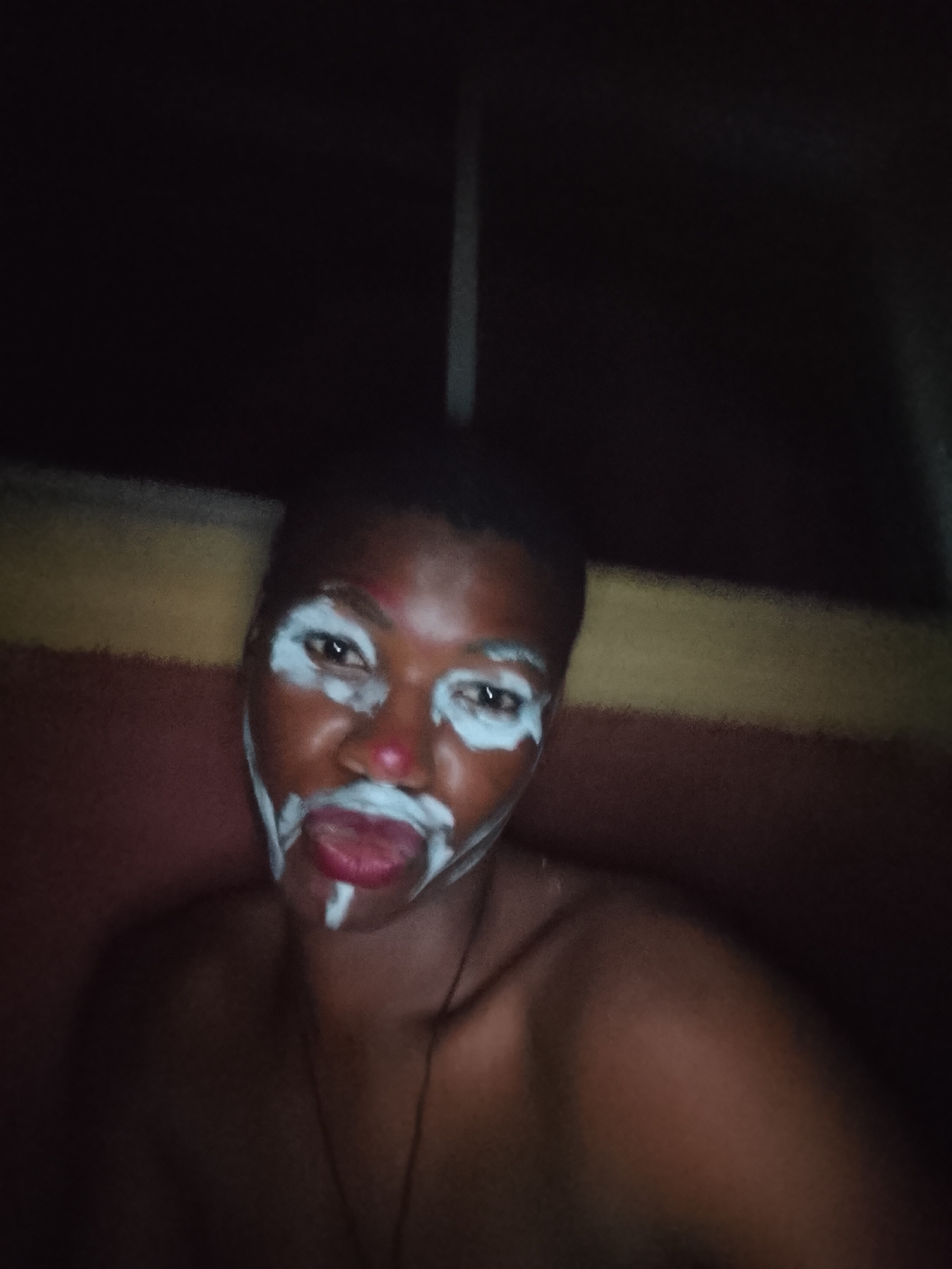 A close-up, poorly lit photograph depicts a person with dark skin and short hair. White paint is applied around the eyes and mouth. The person has their tongue sticking out. A red substance appears to be on the tip of the nose. The individual is unclothed from the shoulders up. A thin necklace is visible. The background is dark, with a horizontal stripe of red, yellow, and brown colors visible at the top behind the person. The location is Unknown, Unknown.