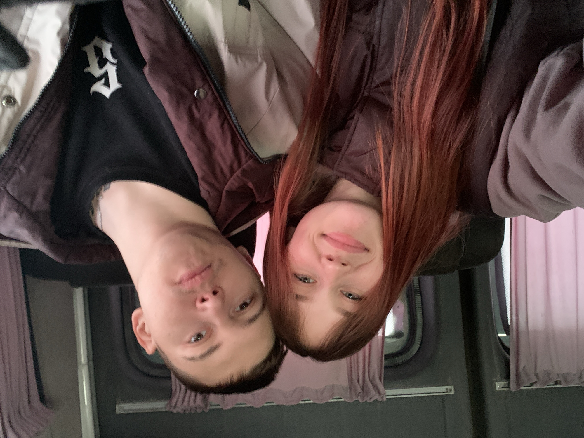 A selfie taken by two individuals. A fair-skinned male with short dark hair is on the left, wearing a black shirt with a white gothic-style letter and a dark brown and beige jacket. To his right is a fair-skinned female with long, reddish-brown hair, wearing a dark brown jacket. The background suggests they are inside a vehicle, possibly a bus or van, with grey walls, windows, and pink curtains. The image is rotated 180 degrees. The location in the city of Votkinsk, Russia, cannot be determined.
