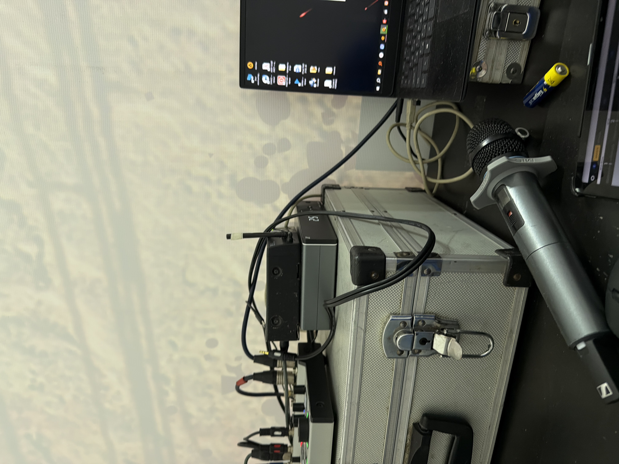 The image shows a collection of electronic equipment on a table. A laptop and a tablet are positioned on the right side of the frame. There is a microphone with a mount placed nearby. The equipment is connected with numerous cables, and power supply units are also visible. The background appears to be a wall with a neutral-colored pattern. The photo was taken indoors in an unknown building or house. The scene gives the impression of an audio-visual setup, potentially for broadcasting, recording, or similar purposes. There is no indication this picture was taken in Perm, Russia.