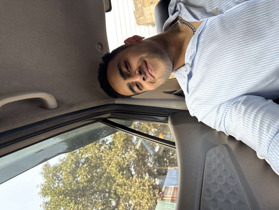 An upside-down image shows a man in a light blue striped shirt, looking toward the camera with a slight smile. The scene is inside a car, evidenced by the interior ceiling, window, grab handle, and gray seat back. Outside the car window, there are trees and some building. The man has short dark hair, and a silver chain is visible around his neck.