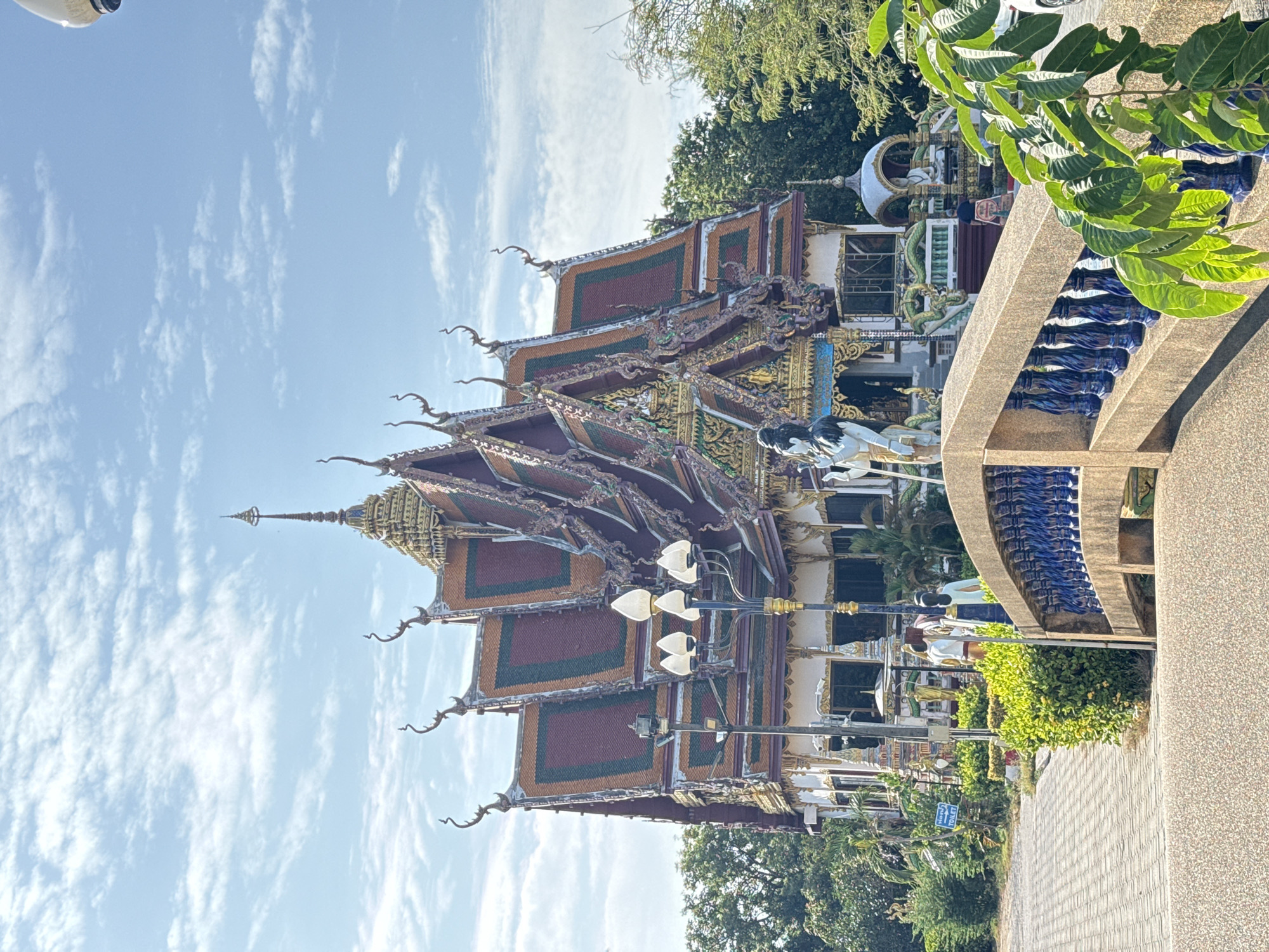 The image depicts a view of a Buddhist temple in Ko Samui District, Thailand. In the foreground, a textured bridge with blue balusters spans across the frame. Beyond the bridge, a multi-tiered temple with ornate, colorful roofing and decorative elements is visible. Several statues are placed around the temple grounds, including a tall, dark-haired figure on the left and a seated Buddha statue under a small dome on the right. Lush green trees and foliage surround the temple. The sky above is a clear blue with scattered white clouds. A paved walkway leads towards the temple entrance. A sign with "TOILET" in English and Thai is positioned near the walkway, indicating visitor facilities.
