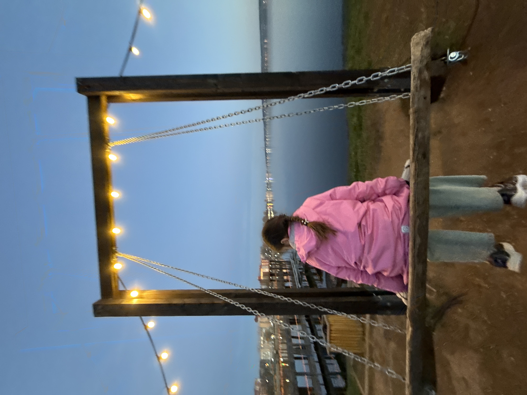 A girl in a pink jacket sits on a swing that appears to overlook the city of Votkinsk, Russia, at dusk. The swing's frame is made of dark wood, decorated with string lights, and hangs by metal chains. In the background, the city lights reflect off the calm waters of what is likely the Votkinsk Reservoir. The surrounding ground appears to be grassy or dirt.