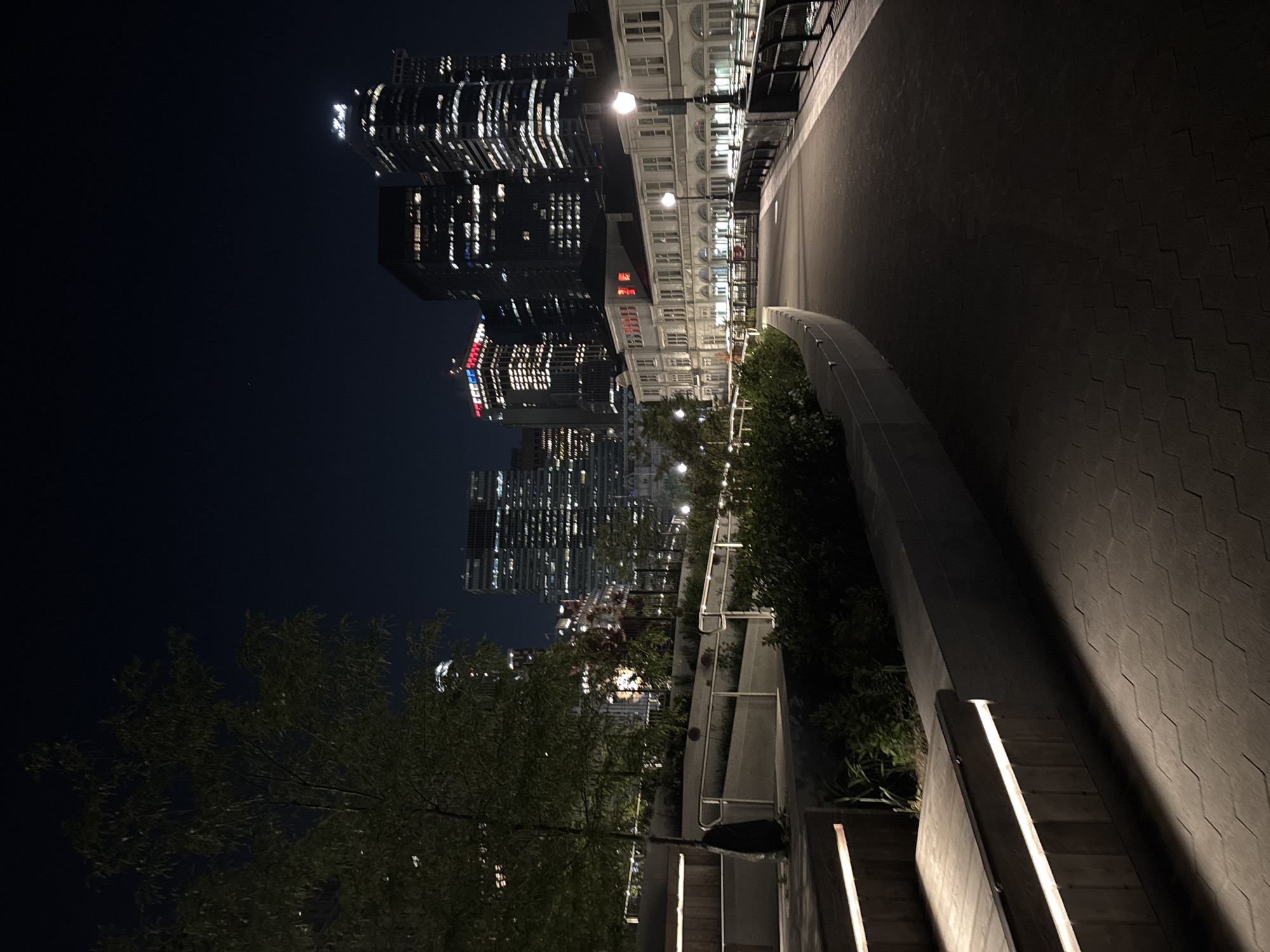 The image presents an urban landscape at night, captured from an elevated viewpoint looking down a modern pedestrian pathway. A dark-tiled walkway, subtly illuminated by integrated strip lighting along a low, light-colored curved wall, extends from the foreground into the distance. To the right of the path, a section of stairs also features embedded strip lighting, casting soft glows on the steps and an adjoining textured wall with a geometric pattern.
On the left side of the path, dense green foliage and trees provide a natural element, partially framing the impressive city skyline. The background is dominated by numerous tall buildings and skyscrapers, their windows brightly lit, suggesting active offices or residences. One prominent modern building displays a distinct red and blue illuminated sign or pattern. Further along the street level, an older, ornate white building with multiple symmetrical windows and visible streetlights stands out amidst the contemporary architecture. The night sky above is dark and clear.
No individuals are visible, creating a sense of quietude despite the bustling city backdrop. The scene evokes a calm yet vibrant urban atmosphere, characteristic of a major metropolis like New York, especially given the context. The advanced lighting design and varied architecture point to a well-developed urban environment. No decipherable text is visible.