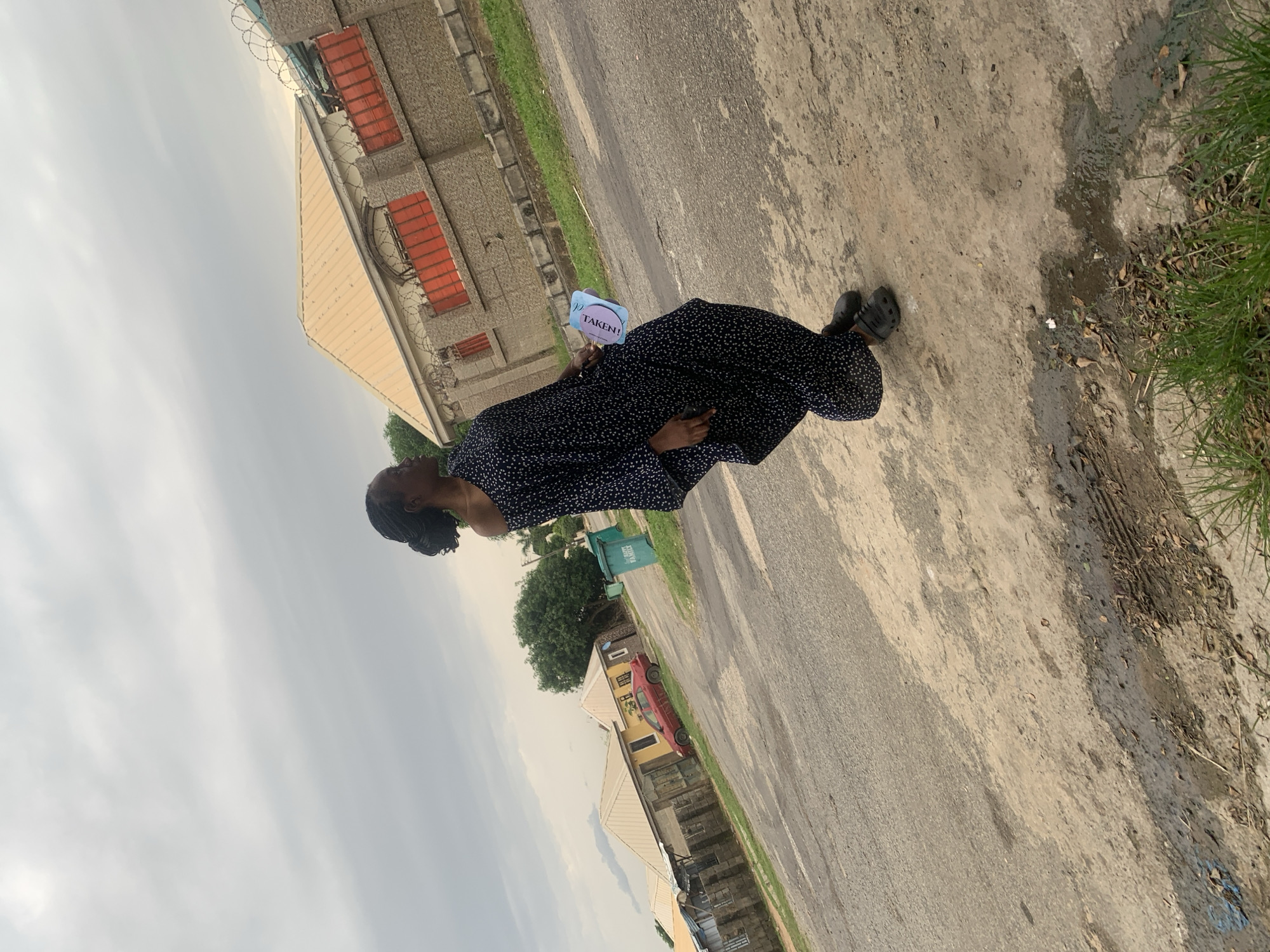 A full-body shot captures a young Black woman standing on a dirt road in Abuja, Nigeria. She is dressed in a long, dark blue, off-the-shoulder dress with white polka dots. Her hair is styled in braids pulled back in a bun. She is wearing dark clogs on her feet. In her right hand, she holds a light blue fan with the word "TAKEN!" printed in purple. To her left, a green trash bin is visible. Behind her, several buildings with orange-tinted roofs line the street. A red car is parked in front of one of the yellow buildings. The sky above is overcast and grey. The road is made of asphalt and dirt, with some puddles and debris scattered on the ground. Green grass grows along the edges of the road.