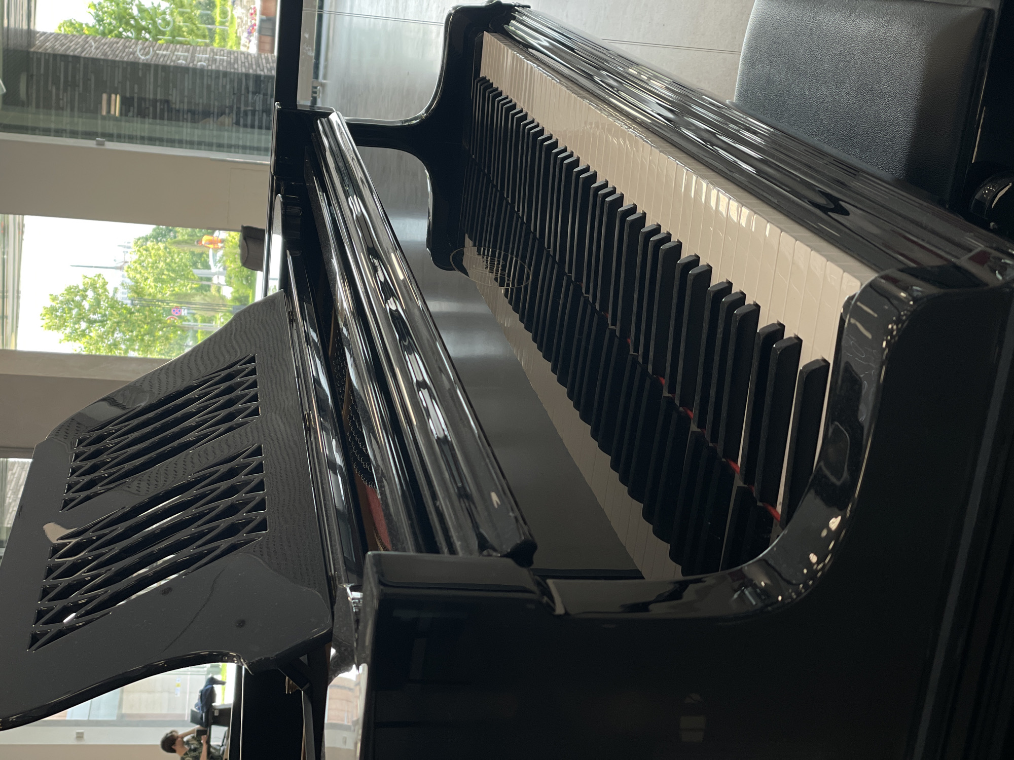 The image shows a close-up view of a black grand piano from a side-angle. The piano is situated indoors, with a large window in the background showing trees and an outdoor environment. The reflection on the piano's glossy surface is prominent. In the background, through the window, a person is visible sitting at a table, and another person is standing outdoors. The scene appears to be in a public space or a building with large windows, possibly a concert hall, lobby, or a music school. The specific location in the city of Kazan, Russia is not evident from the visual information provided in the image.