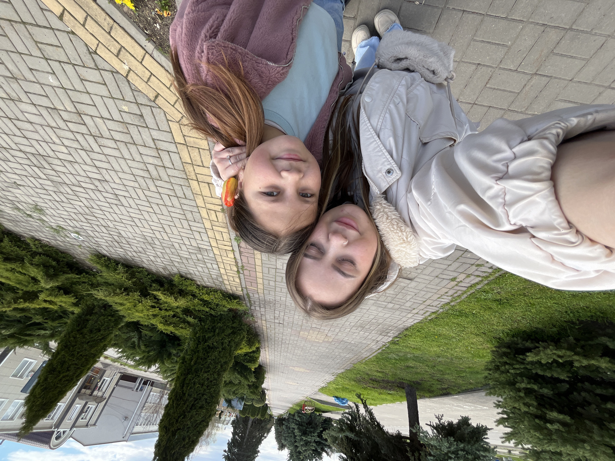 Two young women are pictured outdoors, likely in Novopokrovskaya, Russia. They are positioned head-to-head, close together, suggesting a selfie being taken by one of them. The person on the left wears a pink, fur-like jacket over a light blue shirt, with her brown hair partially visible. She has an object in her hair near her ear. The individual on the right is wearing a light-colored jacket with a fur-trimmed collar. They are on a brick or paved walkway, with a strip of grass and trees visible to the right, and trimmed shrubs and a multi-story building visible to the left. The background shows a typical urban landscape with sidewalks, greenery, and residential structures.