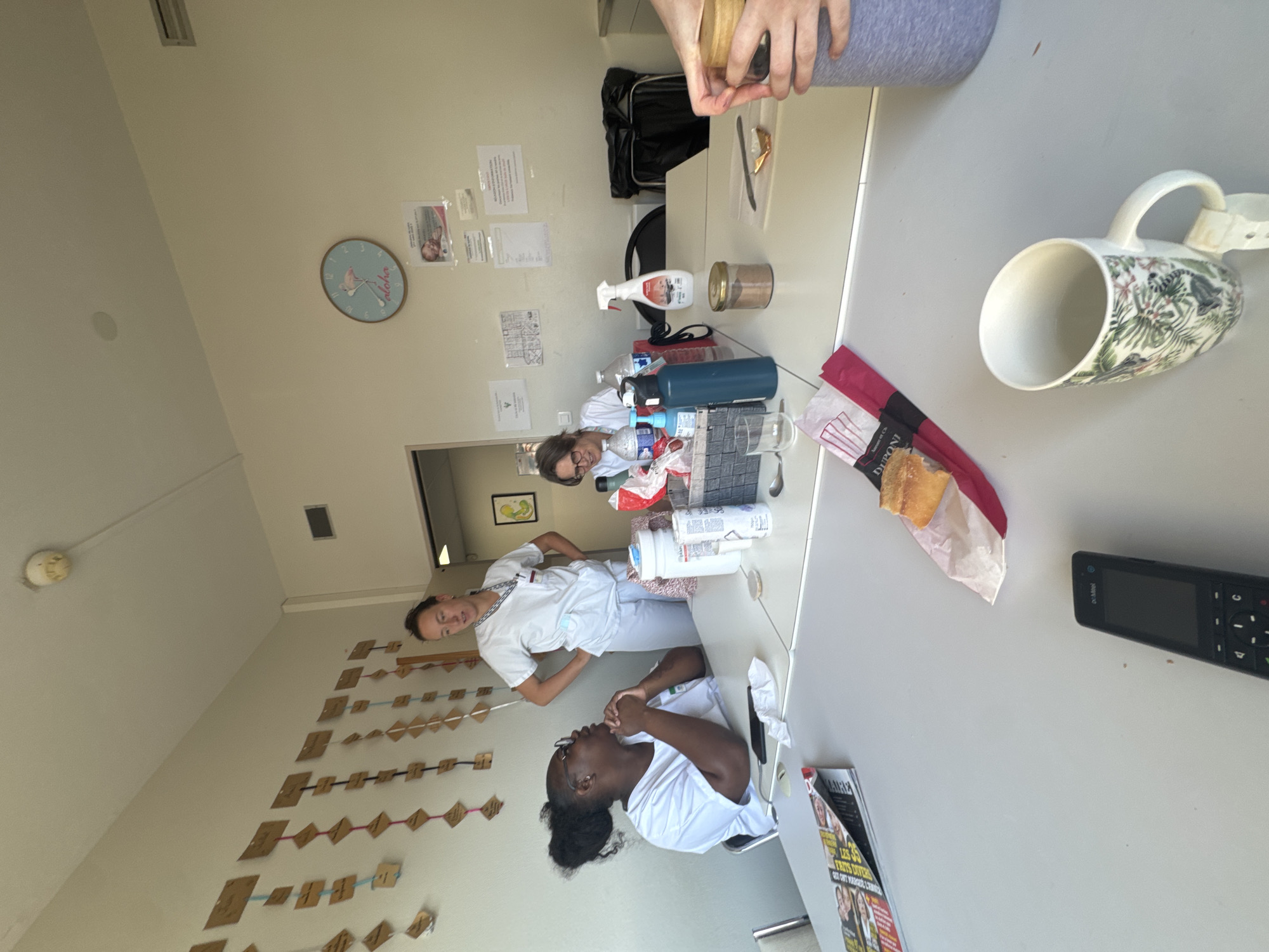 The image depicts an indoor scene, likely a break room or common area, with a focus on a table laden with various items. In the foreground, a white mug with a tropical leaf and monkey pattern sits on the table. To its left, a black cordless phone with a screen is visible. A magazine with headlines in French and images of people is also on the table. A sandwich wrapped in red and white paper lies to the right of the magazine. In the middle ground, two women are seated and one woman stands, all wearing white tops. The seated woman on the left is looking at the standing woman. The standing woman is looking towards the seated woman on the right. The seated woman on the right appears to be looking down and smiling. Several water bottles, a spray bottle, and a container of what appears to be cocoa powder are on the table. A black trash bag is visible in the background. The walls are adorned with numerous small brown tags suspended by colored strings. A clock with a flamingo motif hangs on the wall. The overall impression is a casual gathering during a break. The presence of French magazines and the mention of Tourcoing, France, suggest this location is within that city.