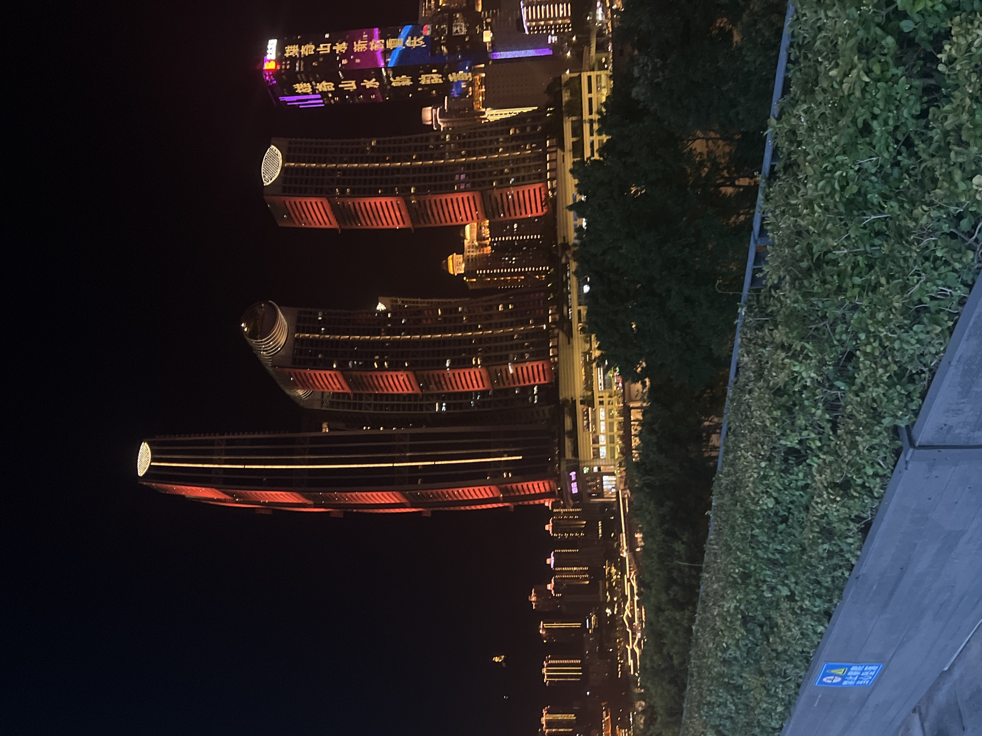 This media file depicts a nighttime view of a cityscape with illuminated skyscrapers and lush greenery. The scene appears to be in an urban environment, possibly in China given the text visible on one of the buildings.
The primary subjects are the modern architectural structures, characterized by their distinctive designs and vibrant lighting. One prominent building features a curved facade with red and yellow lights, giving it a dynamic and futuristic appearance. Another building, further in the background, is adorned with brightly colored lights, including purple and pink, displaying what seems to be text. The text is in Chinese characters and, due to the image's orientation, is difficult to decipher completely. However, some characters suggest it might be related to a commercial or informational display.
The setting is clearly urban at night, with the dark sky contrasting with the illuminated buildings. The presence of dense foliage and what appears to be a paved walkway suggests an area that combines urban development with green spaces, possibly a park or a well-landscaped plaza overlooking the city. The overall mood is one of vibrant urban nightlife, with the lights creating a sense of energy and grandeur.
There are no visible people in the frame, suggesting the photograph was taken from a vantage point overlooking the city. The weather appears clear, as the lights are sharp and there are no signs of precipitation or fog. The time of day is unequivocally night, indicated by the dark sky and the extensive use of artificial lighting. The location context provided, "江北城街道, China," suggests a specific district within a Chinese city, likely a commercial or entertainment hub known for its modern architecture and nighttime illumination. A small blue sign with white text and an icon is visible on the wooden structure on the right side of the frame, but the text is too small to read.