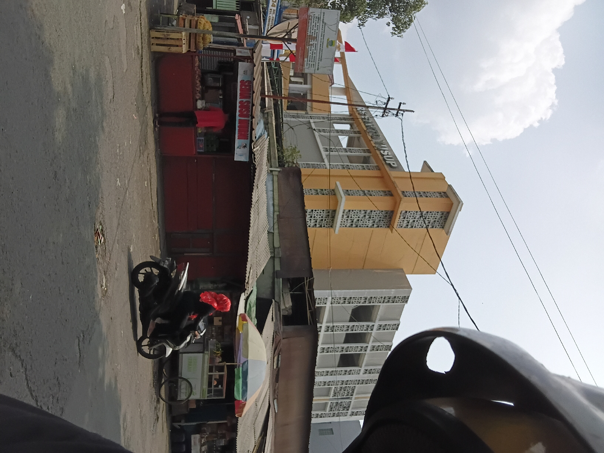 The image captures a street scene in Bandung City, Indonesia, on a cloudy day. In the background, a large, multi-story building with orange and white sections stands prominently. The building has a distinctive architectural style with patterned concrete blocks. To the left of this building, a signpost displays information, including what appears to be hospital or institutional branding with Indonesian text and red and white flags.
In the mid-ground, a row of low-rise buildings with corrugated metal roofs lines the street. One of these buildings, painted red, has a sign that reads "SEDIA SUSU MURNI," suggesting it's a stall selling fresh milk. A person wearing a red shirt is visible behind the counter of this stall.
In the foreground, a woman wearing a red headscarf and dark clothing is riding a black and white motorcycle. She is positioned on the right side of the frame, heading towards the viewer's right. The road surface is asphalt with visible cracks and patches. To the far right of the image, the edge of a dark-colored helmet, likely from the perspective of the photographer, is visible. Behind the woman on the motorcycle, a stall with a colorful umbrella is set up, and a cart with wheels is parked nearby. Trees are visible on the far left of the image. The overall impression is that of a busy urban street with commercial activity.