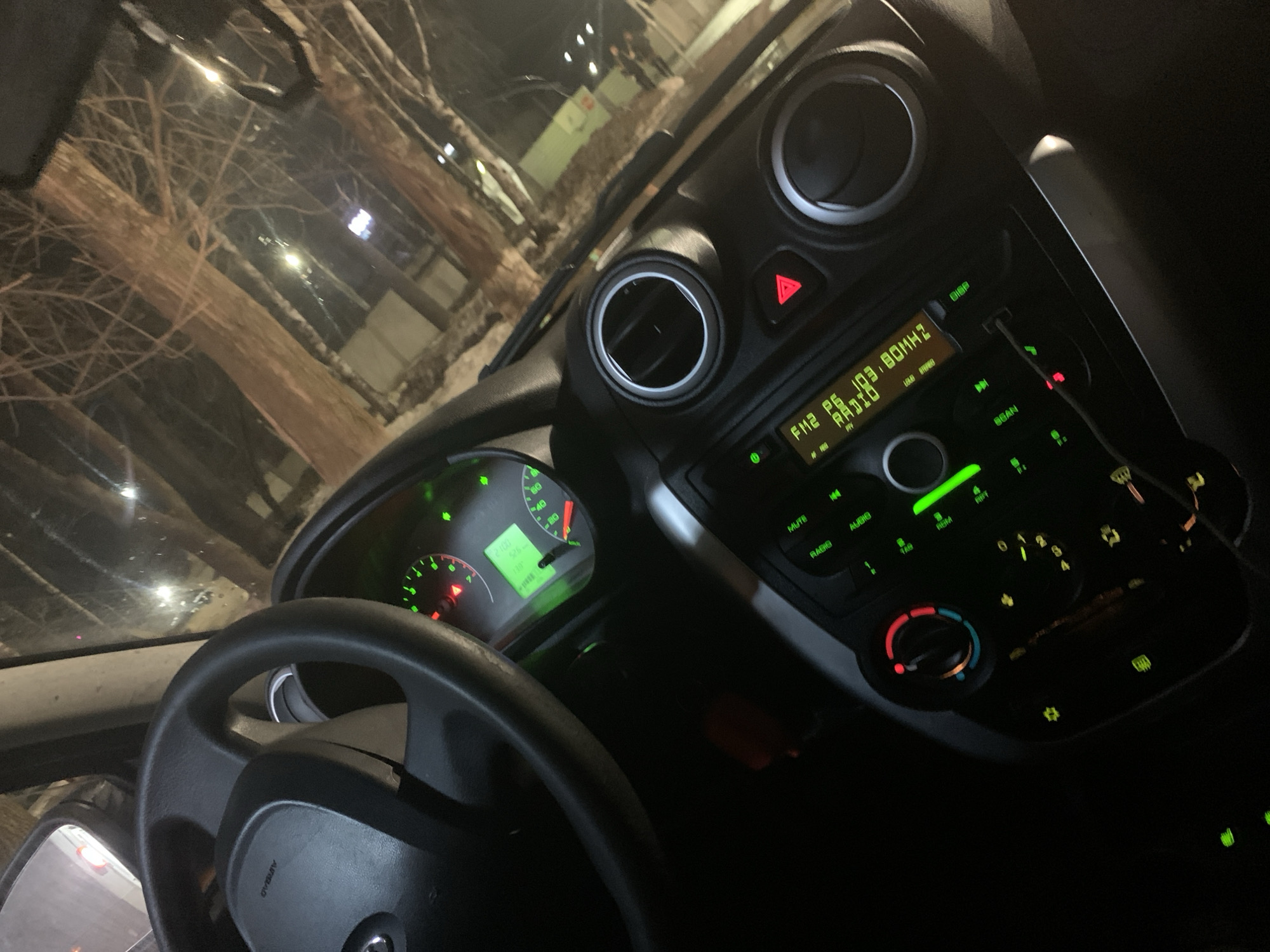 The image shows the interior of a car, focusing on the dashboard, steering wheel, and windshield view. The car appears to be stationary at night.
Dashboard: The instrument panel is illuminated with green lights. The speedometer reads approximately 20 km/h. The radio display shows "FM2 P6 103.80MHZ RADIO". Other controls are visible, including buttons for "MUTE," "RADIO," "AUDIO," "SCAN," and climate control settings with red and blue indicators.
Steering Wheel: A black steering wheel with a center emblem is in the lower left of the frame.
Windshield View: The view through the windshield shows a blurred urban landscape at night. Tree branches are visible, along with streetlights.
General: The overall lighting is dim, with the dashboard lights being the primary source of illumination. The scene suggests the car is parked or idling at night. No people are visible inside the car.