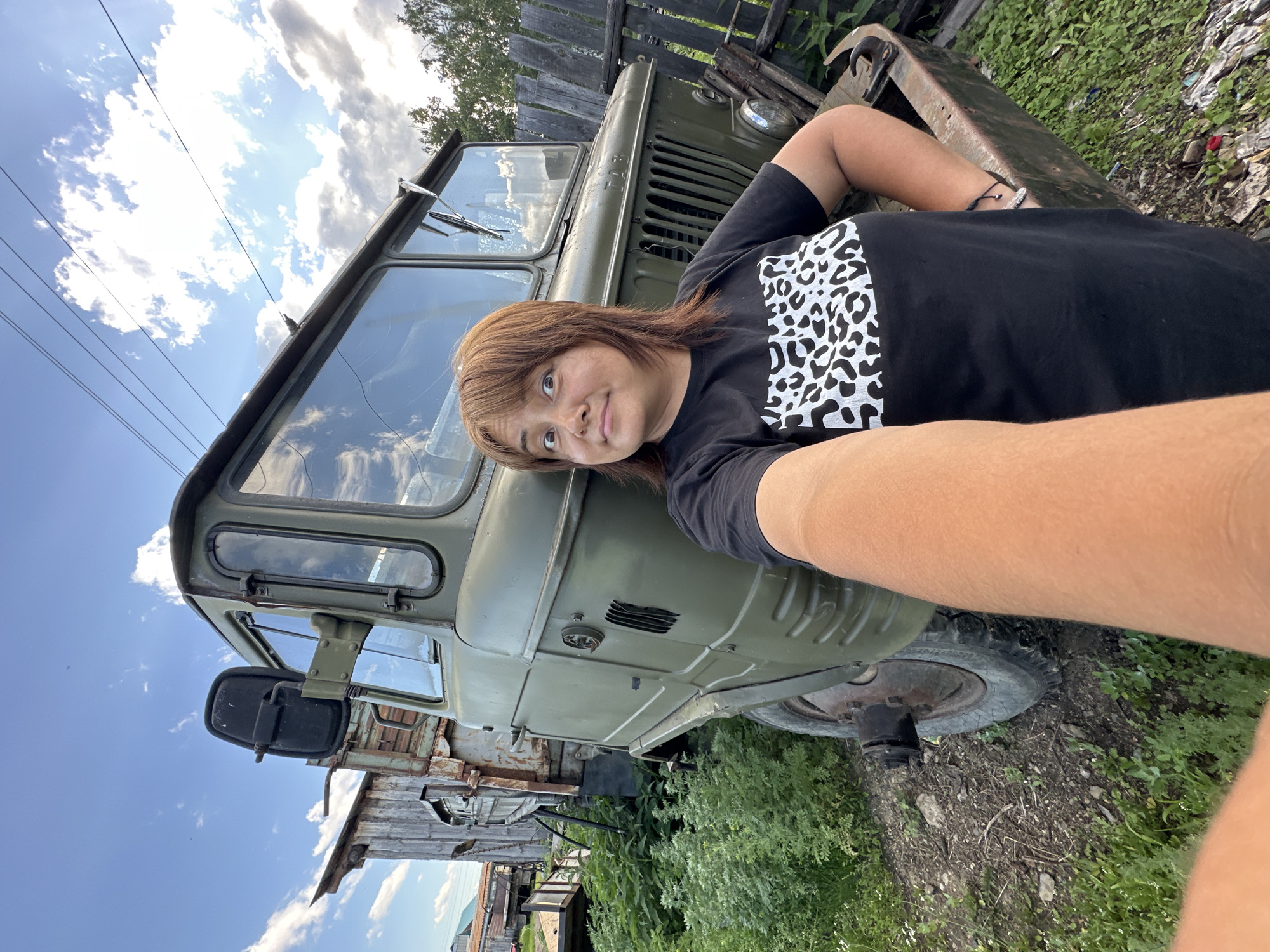 A person is lying on the hood of an old, olive-drab military truck, taking a selfie. The person has shoulder-length brown hair and is wearing a black t-shirt with a white leopard print design on the chest. Their face is turned towards the camera, with a slight smile and a direct gaze. The truck appears worn and weathered, with rust visible on some parts. The scene is outdoors, with green foliage and a wooden fence in the background. The sky is bright blue with scattered white clouds, suggesting it is daytime and the weather is fair. The overall impression is casual and relaxed, with the person enjoying a moment outdoors next to the truck. There is no visible text in the image.