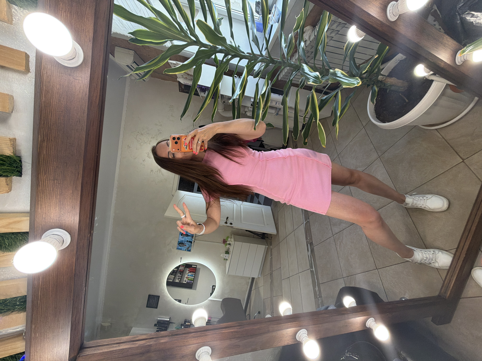 A young woman is posing for a selfie in front of a large vanity mirror. She is wearing a pink dress and white sneakers. She is holding a smartphone in her right hand and making a peace sign with her left hand. The mirror is surrounded by light bulbs. The reflection in the mirror shows a room with a round mirror, a chair, cabinets, and a plant. The location may be Birobidzhan, Russia, given the reflection in the mirror shows an interior of a beauty salon or hairdresser shop.