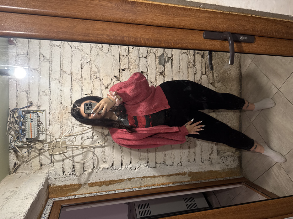A female subject is depicted in a mirror selfie taken within an interior space in Pryaporets, Bulgaria. She stands centrally, utilizing a smartphone to obscure her face. She is dressed in a pink knitted open cardigan, a dark t-shirt featuring a red lip graphic, black trousers, and light grey socks. Her visible accessories include a silver cross necklace, extended white artificial fingernails, and a light pink scrunchie on her right wrist. Script tattoos are visible on her left ankle and right forearm.
The background features an unpainted white brick wall. An exposed electrical service panel, characterized by a complex array of visible wiring, is mounted on the upper left section of this wall. A single bare light bulb is affixed to the wall above the electrical panel. To the left, a light-colored wooden door frame with a glass panel provides a partial view into an adjacent room, where a white, multi-vented appliance is visible. The floor is covered with light-colored ceramic tiles. To the right, a dark wooden door with a dark handle and lock serves as the reflective surface for the photograph.