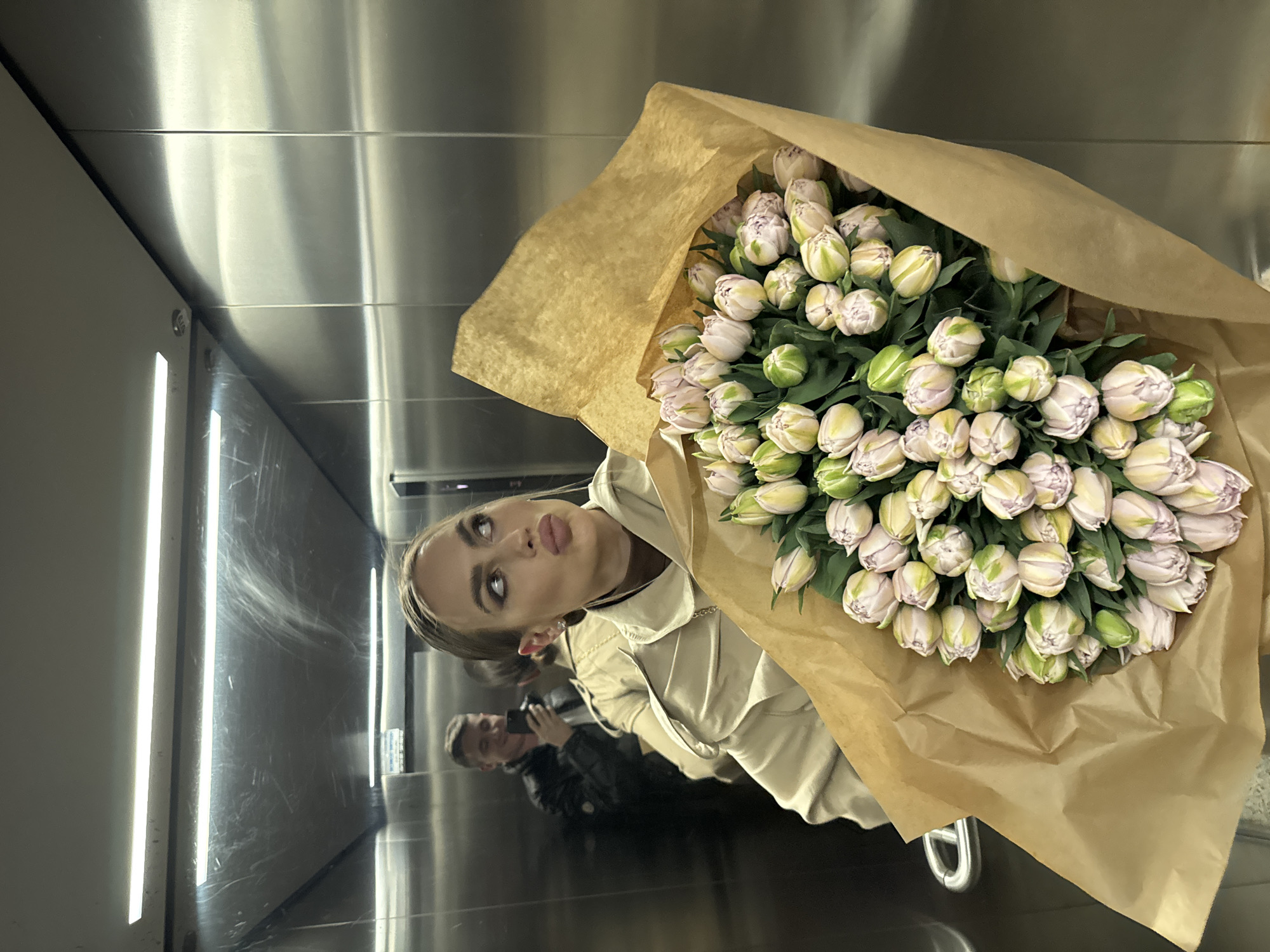 A person is standing in an elevator in Warsaw, Poland, holding a large bouquet of light pink and green tulips wrapped in brown paper. The walls and door of the elevator are made of a reflective material. The person is wearing a light-colored jacket and has their hair pulled back. Another person can be seen reflected in the elevator's mirrored surface, holding a phone as if taking a picture. The fluorescent lights are visible on the left side of the elevator's interior.
