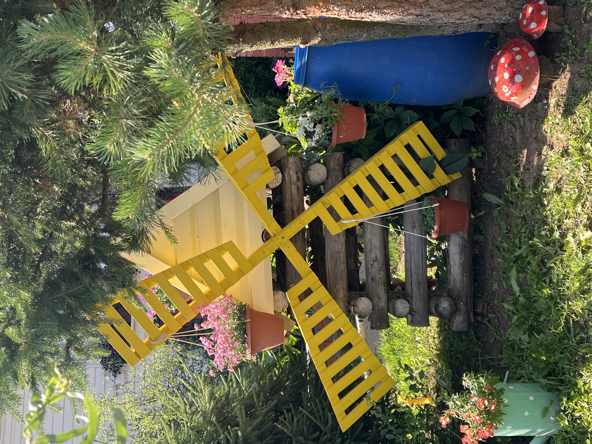 The image depicts a decorative garden scene in the city of Perevoznoe, Russia. In the foreground, two red and white toadstool garden decorations are visible on the ground. To the left of the toadstools, a green watering can with white polka dots holds an arrangement of red flowers. Behind the watering can, a rustic yellow windmill decoration is the central focus. The windmill is constructed from wooden planks and rests on a base of stacked logs. Two potted plants hang from the windmill structure: one with pink flowers above the windmill blades, and another with white and green foliage to the right. Behind the windmill and to its right, a large blue barrel is partially visible, next to a tree trunk with rough bark. The scene is set outdoors, with lush green grass and various plants and trees forming the background. The lighting suggests it is daytime, with sunlight filtering through the leaves of a large conifer tree overhead, casting shadows on the ground.