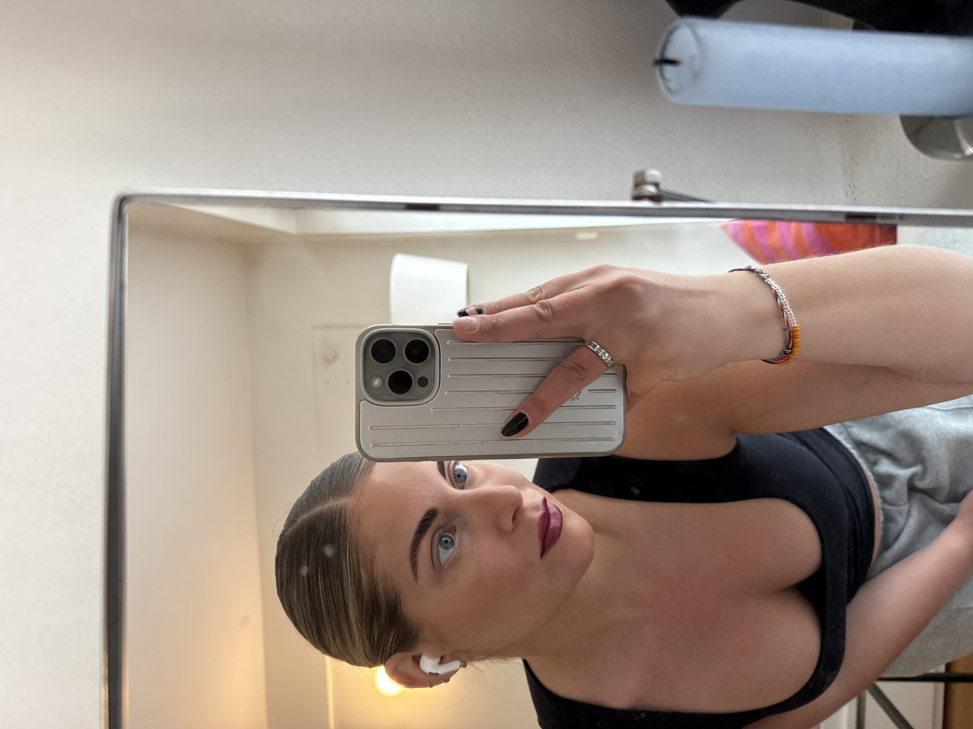 A woman takes a selfie in a mirror. She is wearing a black tank top and gray sweatpants. She is holding an iPhone in her right hand. She has dark hair pulled back and is wearing an AirPod in her left ear. She has a bracelet on her right wrist and a ring on her right hand. The background is a white wall and a sink. The reflection in the mirror shows a room with white walls. It is not possible to determine the location in Paris from this image.