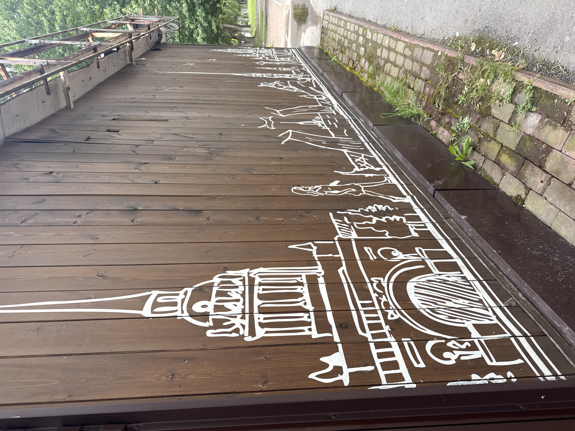 The image depicts a wooden wall adorned with a white line drawing of a city skyline. The drawing features several buildings, including a prominent one with a spire, arches, and columns, reminiscent of classical architecture. A silhouetted figure of a person walking is also visible. The wall itself is constructed from vertical wooden planks and is painted a dark brown. The artwork extends horizontally across the wall. At the bottom of the wall, there is a dark brown base. In front of the wall, a cobblestone path is laid, with some patches of grass and weeds growing between the stones and along the edge. A wet asphalt surface is visible to the right of the path, suggesting recent rain. In the background, trees and foliage are present, indicating an outdoor setting.
The drawing style is simple and somewhat abstract, with clean white lines defining the architectural forms and the human figure. The overall impression is that of a mural or graffiti art on an exterior surface. The context of Ufa, Russia, is not visually evident from the image itself but is provided as information about the location. Therefore, it's not possible to confirm or deny the specific location within Ufa based solely on the visual content.
