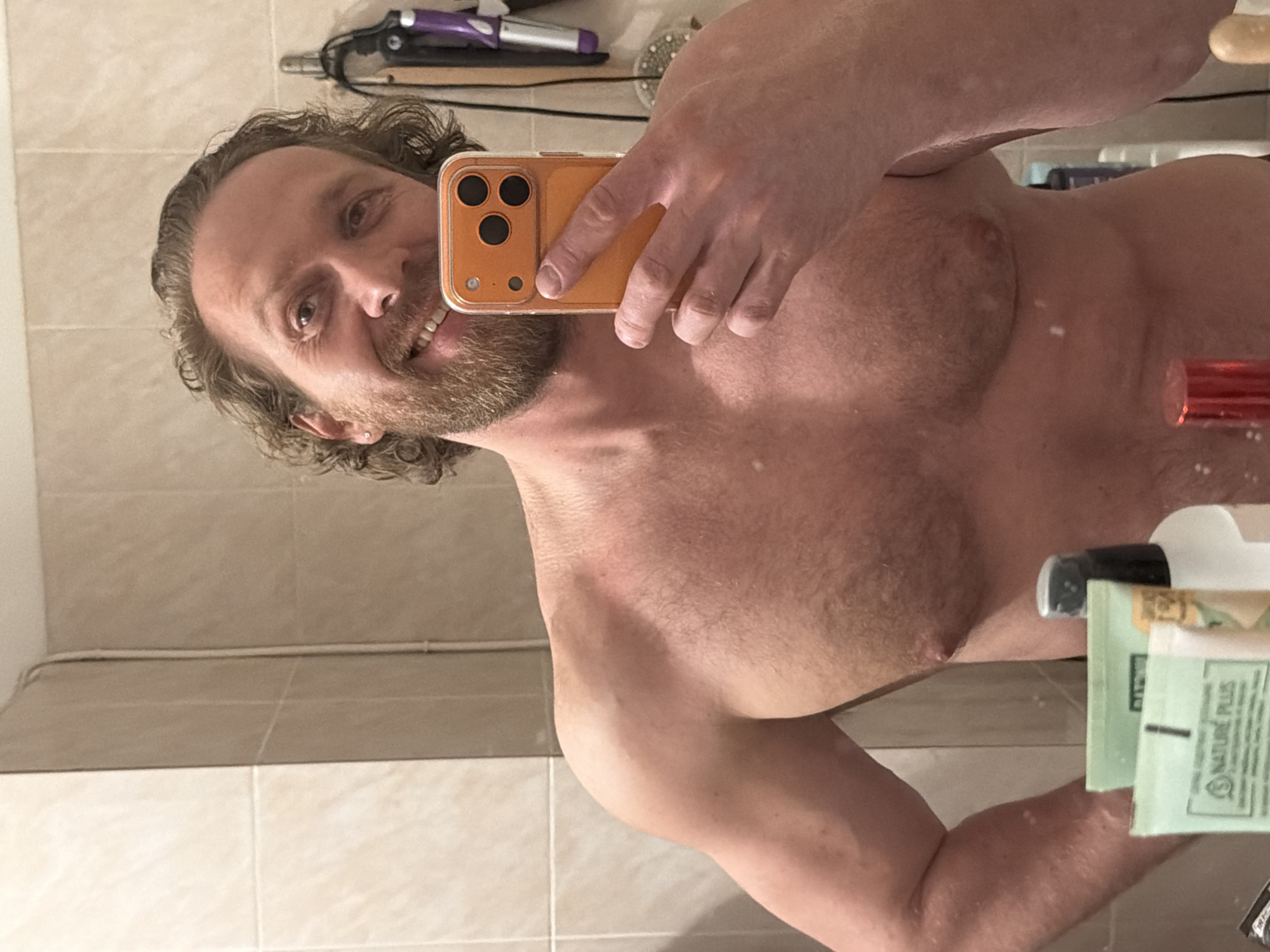 A man with brown hair and a beard is taking a selfie in a bathroom mirror. He is shirtless, and his upper body is visible. He is holding a smartphone with an orange case in his right hand, which is partially obscuring his face. In the background, there is a tiled wall and a shelf with various items, including what appears to be a hair styling tool and several bottles of toiletries. The scene does not provide sufficient information to identify the location as Saint Petersburg, Russia.