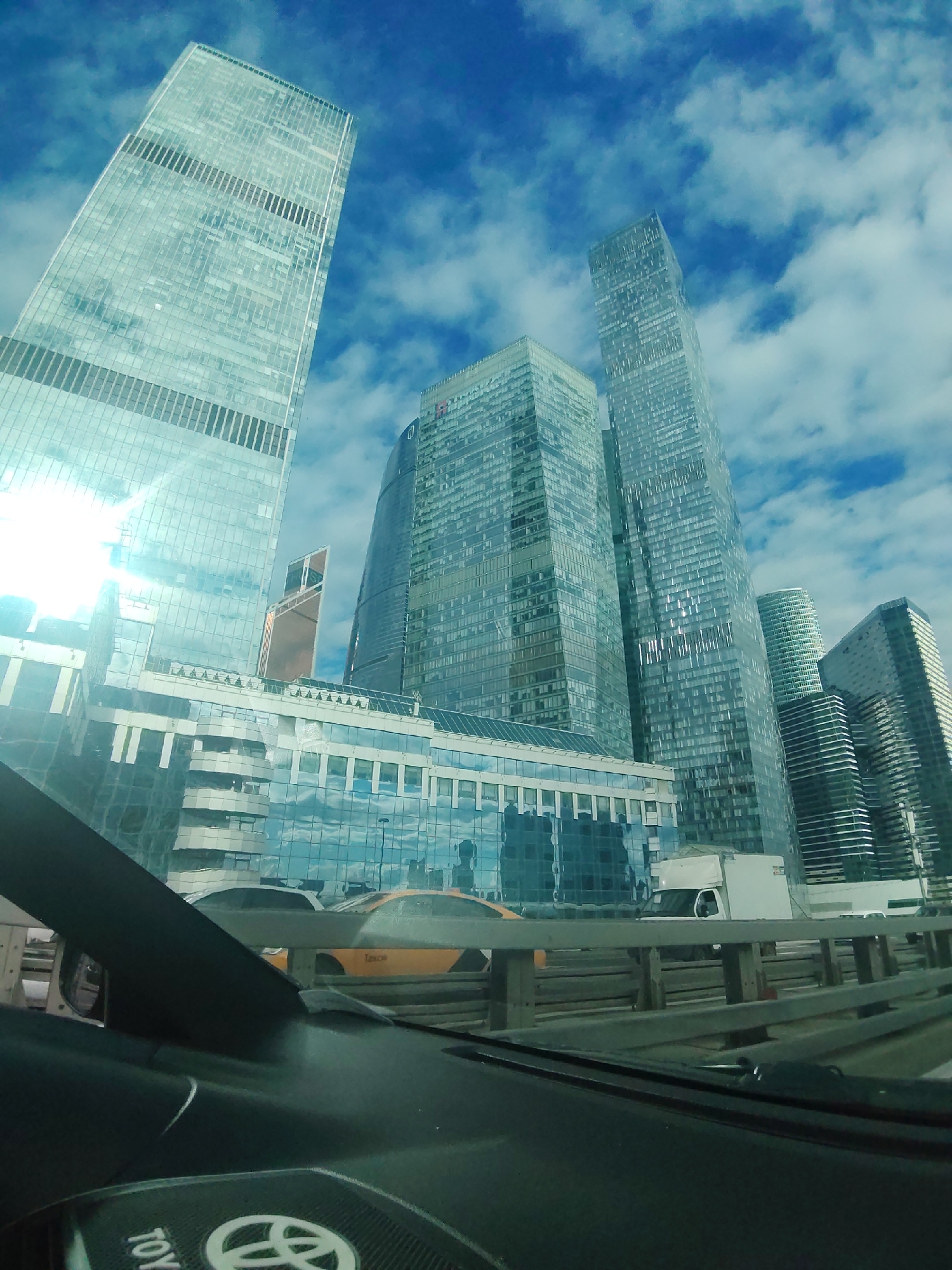 The image shows a view from inside a vehicle looking out at a collection of modern skyscrapers in Moscow, Russia. The buildings are tall and made of glass, reflecting the cloudy blue sky. The closest building in the foreground is a lower structure with multiple stories and glass windows. In the middle ground, several very tall skyscrapers dominate the skyline, their facades appearing to be made of numerous glass panels. To the right, other tall buildings are visible, some with curved or angular designs. In the lower part of the image, the interior of a vehicle is visible, including a dashboard and part of a steering wheel with a Toyota logo. In the scene outside the vehicle, there are yellow taxis and white trucks on a road or highway. The overall impression is of a busy urban environment with contemporary architecture under a partly cloudy sky.