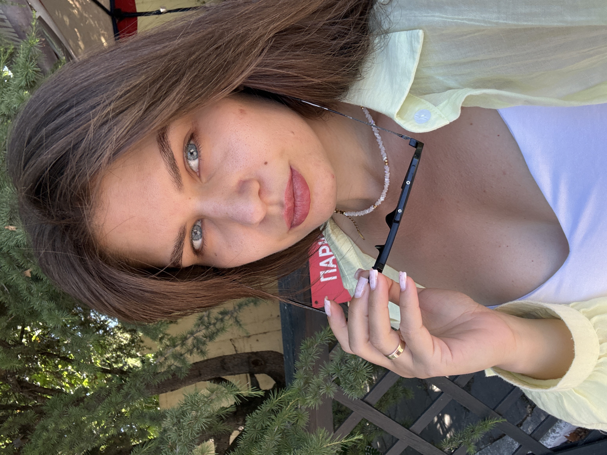 A young woman with long brown hair and blue eyes is holding a pair of glasses in her right hand, her fingers lightly gripping the arms. She is wearing a pale yellow collared shirt over a white top, and a beaded necklace around her neck. A gold ring is visible on her left ring finger. In the background, there is lush green foliage and a blurred view of an outdoor seating area with a striped umbrella. A red sign with white Cyrillic lettering, reading "ПАРК," is visible behind her. The scene appears to be outdoors on a sunny day.