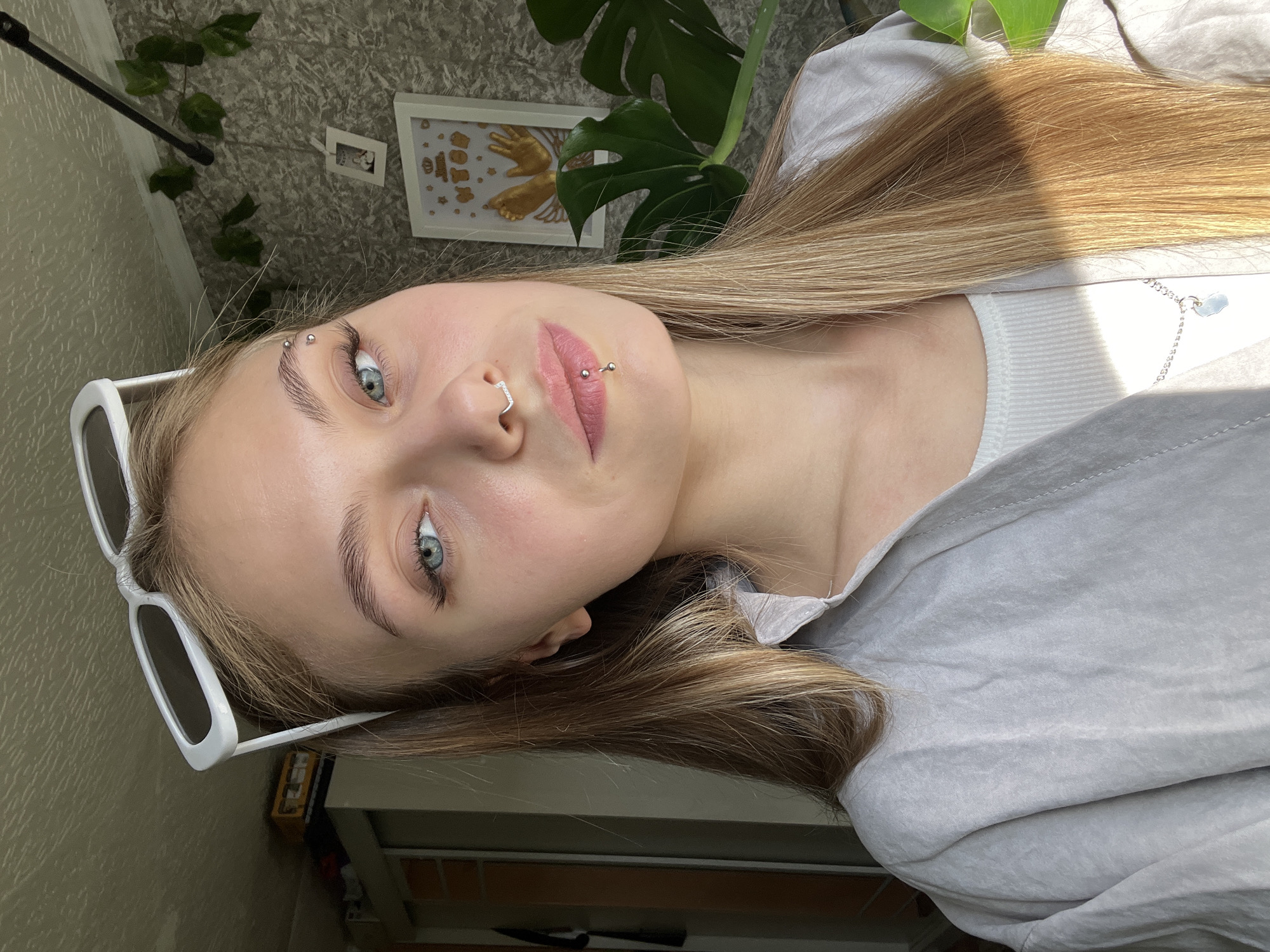 A young woman with blonde hair is posing for a selfie indoors. She is wearing white sunglasses and a light-colored top. She has piercings on her eyebrow, nose, and lip. Behind her is a plant with large leaves and a framed picture on the wall. The location is not discernible from the image to determine if it is in Фоки, Russia.