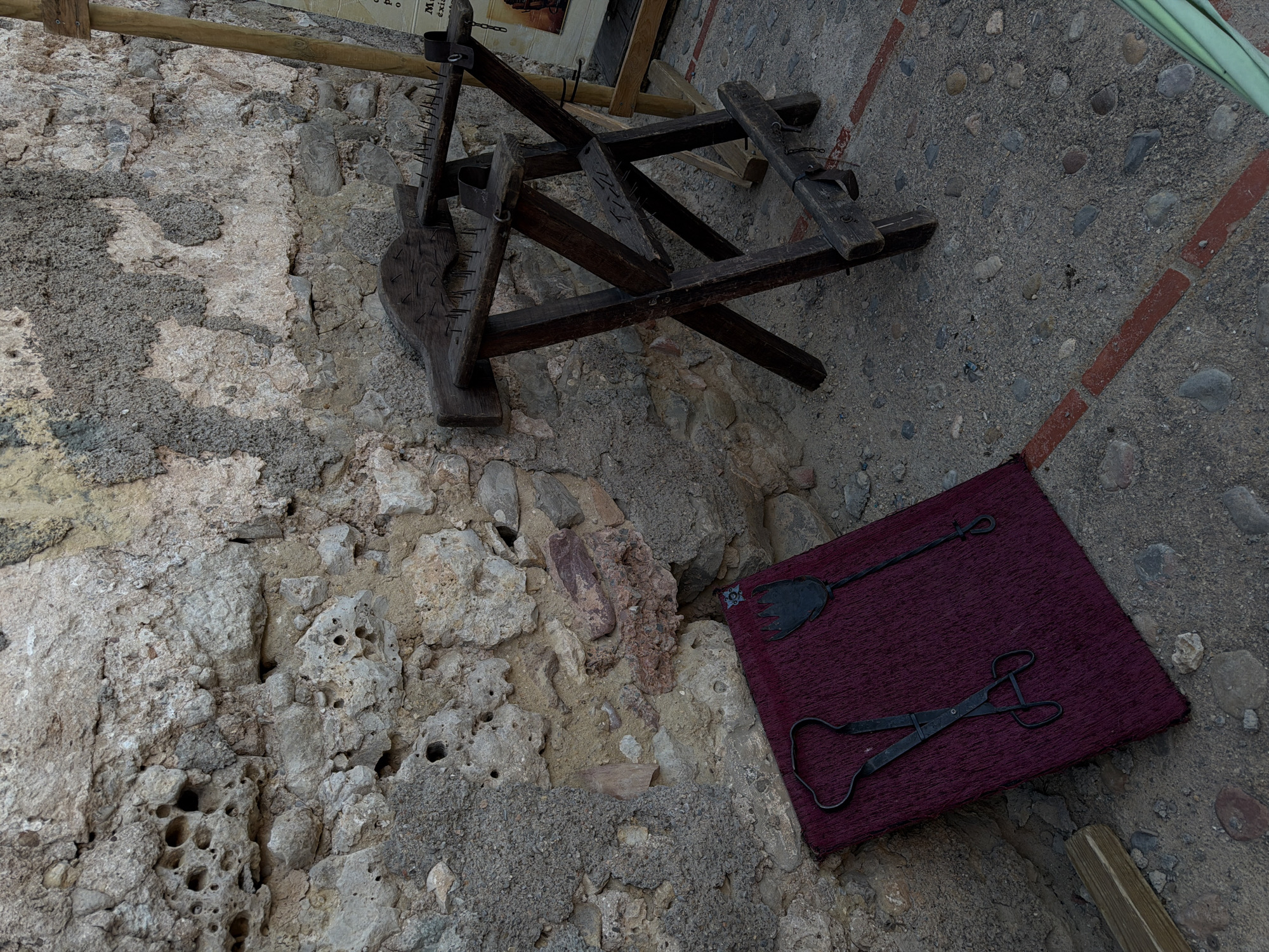 The image shows a high-angle view of a scene likely staged for historical display in Burjassot, Spain. On the left, there's a textured stone wall with varying shades of grey and beige, exhibiting a weathered appearance. Above it is what appears to be a wooden torture device with visible spikes, perhaps a rack or similar instrument. To the right of the torture device is a concrete slab section with red bricks bordering a corner. A small purple fabric mat rests on the stone slab, displaying two vintage iron tools: a flat-bladed spade and a pair of tongs. The overall composition suggests an intentional arrangement for exhibit or photographic purposes, evoking a sense of history or medieval times.