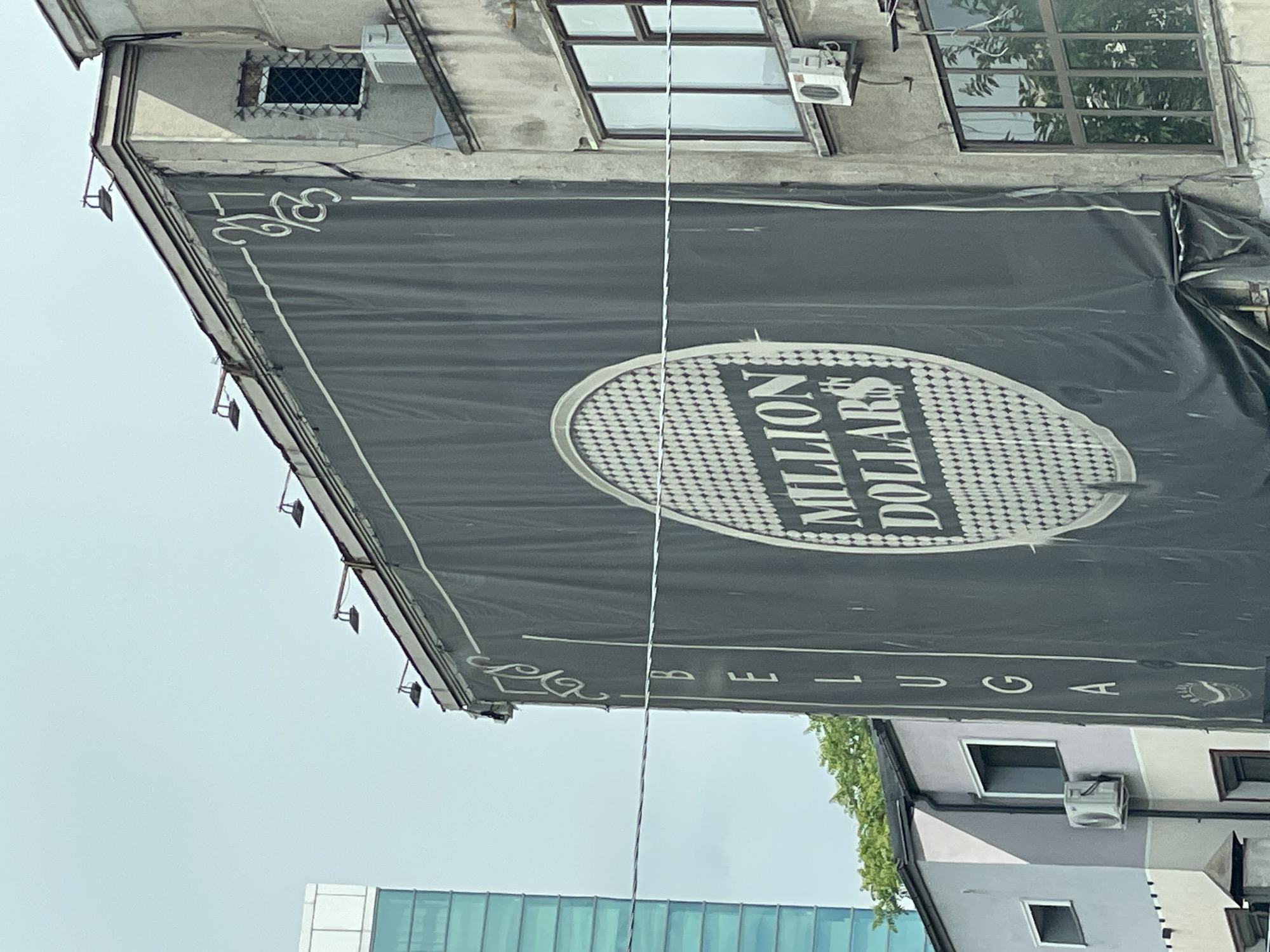 A billboard for "Million Dollars Club Beluga" is mounted on a building in Bucharest, Romania. The sign is primarily black with white lettering and trim. Above the billboard are windows, some with air conditioning units. Below, parts of other buildings are visible, including a modern glass structure and a light-colored building. The sky is overcast.