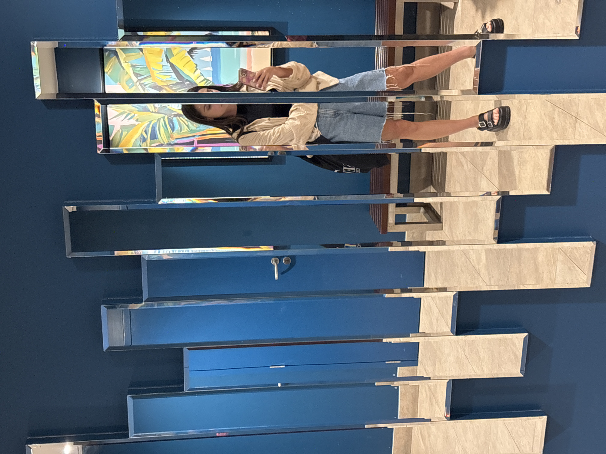 The image shows a young woman taking a mirror selfie in a public restroom. The restroom walls are painted a dark blue, and there are multiple vertical mirrors of varying lengths on the wall. The woman is wearing a black t-shirt, a light-colored open jacket, and denim shorts. She is holding a pink smartphone in her right hand and is looking at its screen. She is wearing black platform sandals. In the background, through the mirrors, a colorful painting of tropical plants and sailboats can be seen, as well as a wooden bench. The floor is tiled in a light beige color. The woman's reflection is visible in the mirrors, showing her from the chest down. The image appears to have been taken in Amman, Jordan, likely in a modern establishment such as a restaurant or shopping mall.