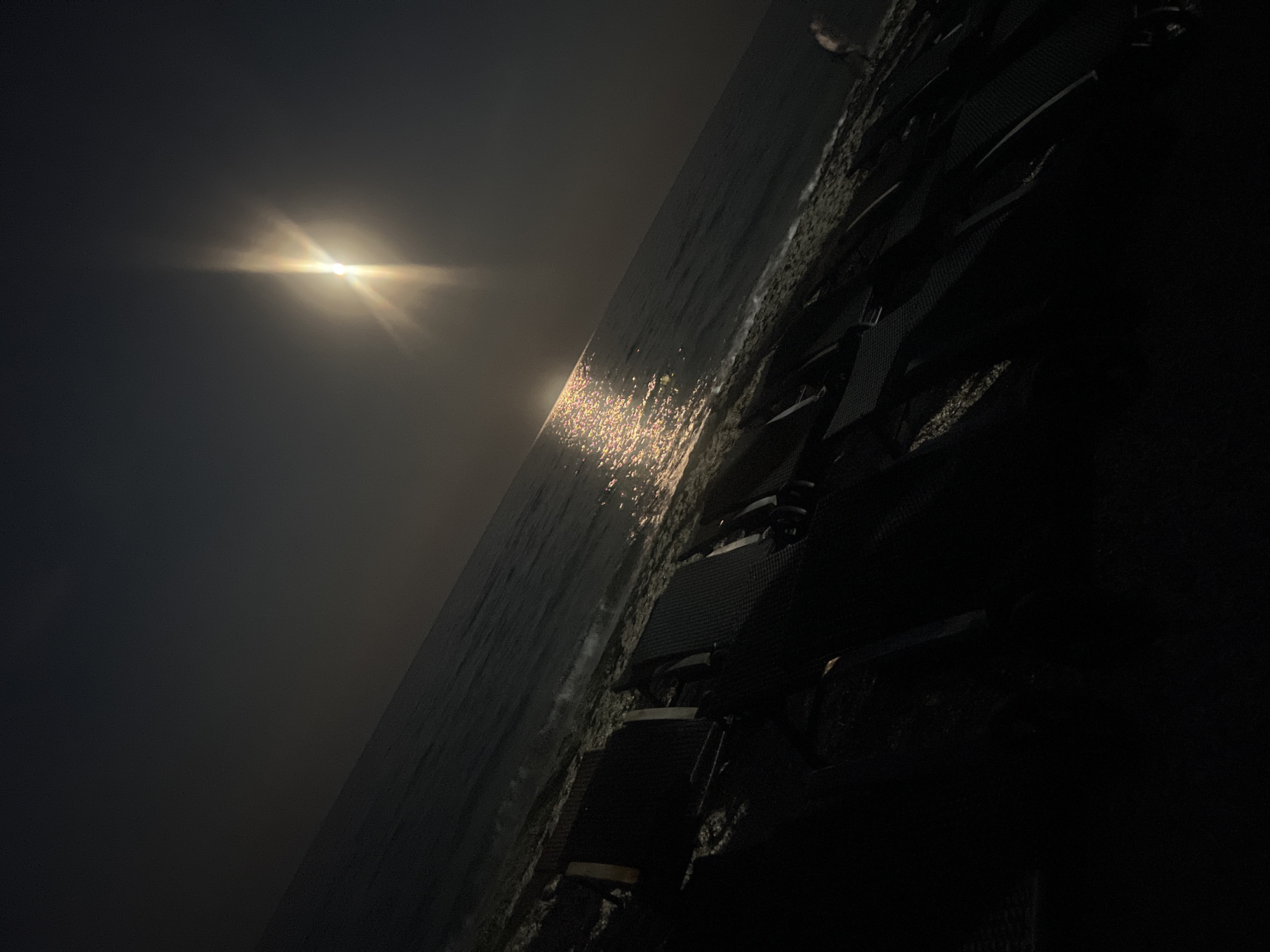 The image depicts a dimly lit beach scene with a body of water in the background, likely the Black Sea, given the location of Odesa, Ukraine. The sun, low on the horizon, casts a brilliant glare on the water, creating a shimmering effect. In the foreground, rows of empty lounge chairs are arranged on the dark sand, suggesting a beach resort or public area. The lighting is subdued, with the sun being the primary light source, creating strong contrasts and shadows. The overall atmosphere is one of tranquility and possibly early morning or late evening on the coast.