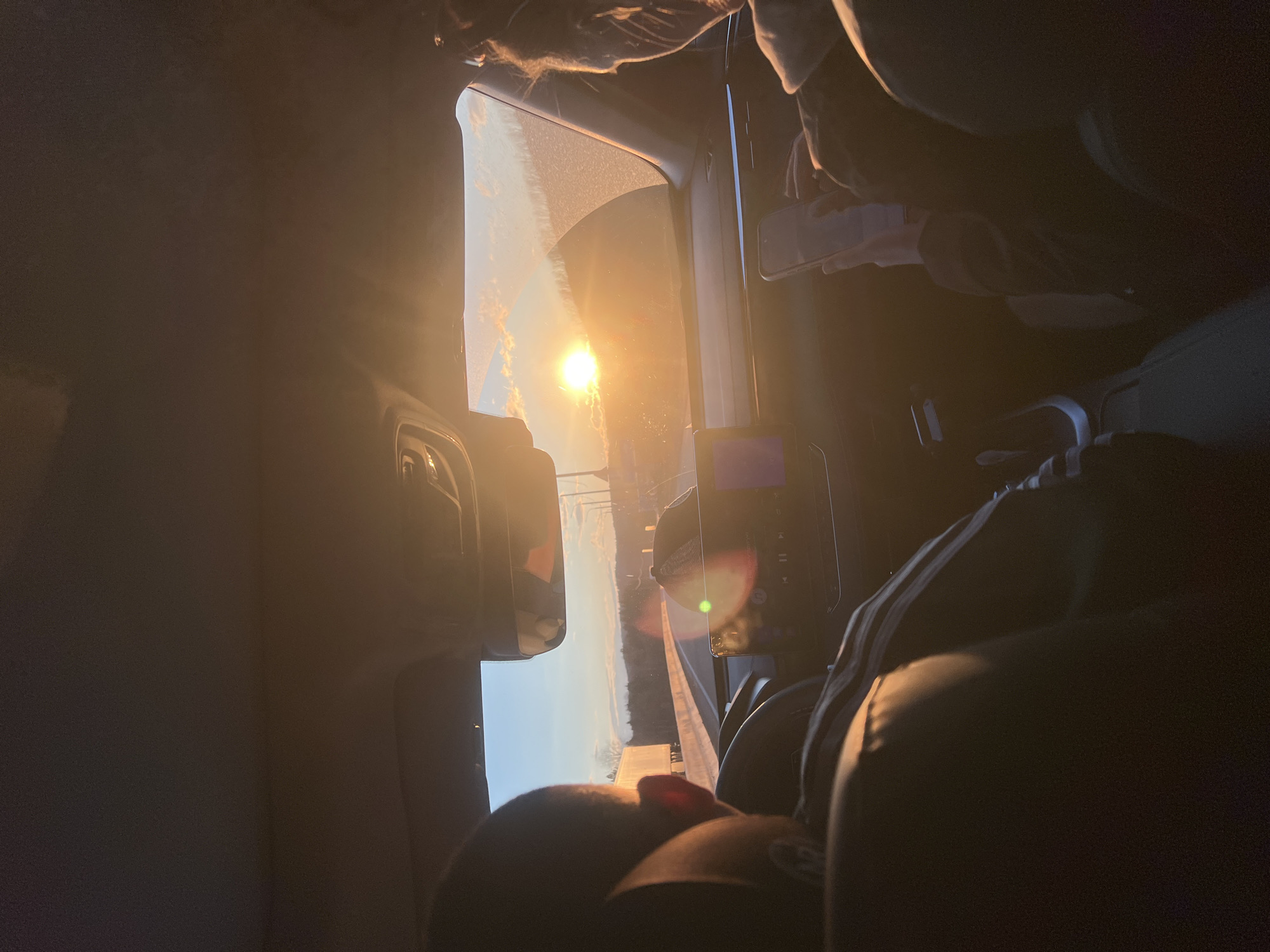 This image captures the interior of a vehicle, likely a car or bus, during either sunrise or sunset, with a prominent sun casting a warm, golden glow. The photograph is rotated 90 degrees counter-clockwise.
In the foreground, a person is visible from behind, resting in a seat, with their head and shoulders partially obscured. They are wearing a dark baseball cap. Further back in the vehicle, other passengers are indistinctly visible, mostly as dark silhouettes or partial limbs (e.g., legs).
The bright sun, low on the horizon, is the dominant feature, shining intensely through a side window and creating significant lens flare. Its rays illuminate dust or condensation on the window pane. Outside the window, a clear sky with a distant horizon and what appears to be a road or open landscape can be seen. Inside the vehicle, parts of the dashboard, a side mirror, and a small electronic screen (unreadable due to distance and glare) are visible. The entire scene is bathed in a soft, diffused light, highlighting the peaceful, possibly sleepy, atmosphere of travel.
No specific text is discernible on any objects within the image. The time of day is clearly morning or evening due to the sun's position and the quality of light. The specific location of Tiush, Russia, is not identifiable from the visual content.