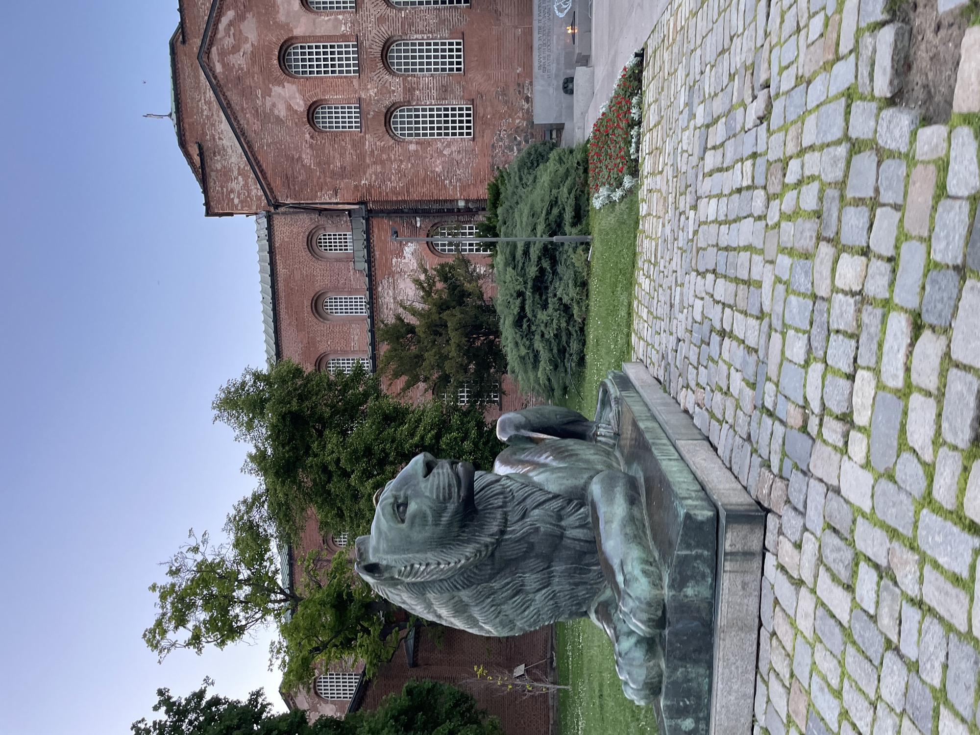 The image shows a bronze lion statue in Sofia, Bulgaria. The statue is situated on a stone base next to a cobblestone walkway. Behind the statue, there is a red brick building with arched windows, interspersed with trees and shrubbery. The sky is visible in the upper left corner and is light blue. Part of the monument in front of the Unknown Soldier can be seen behind some greenery.
