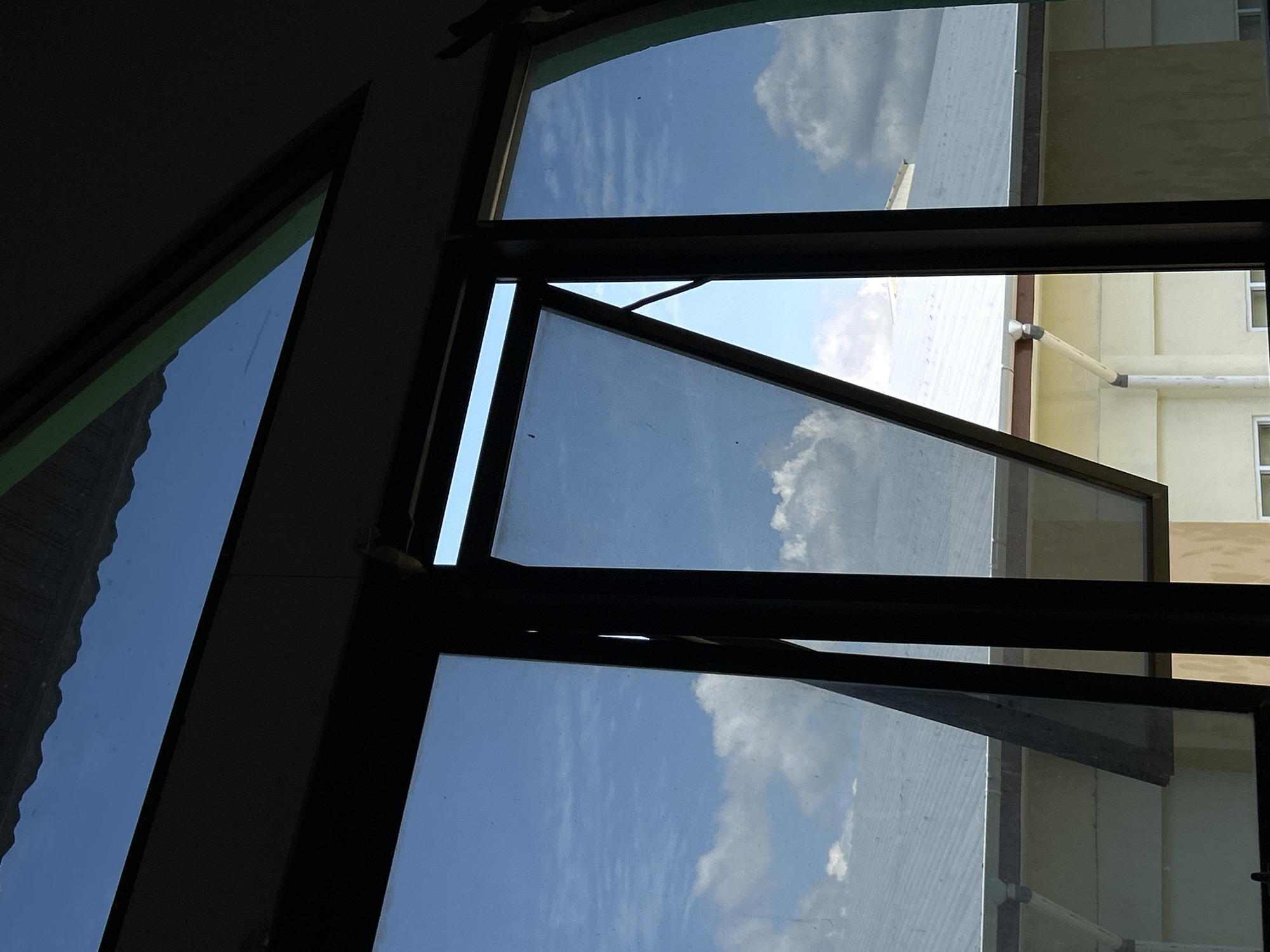 The image provides an indoor perspective looking out through a series of windows. The view is primarily composed of dark window frames that divide the scene into multiple glass panes. Many of these panes are closed, reflecting a bright blue sky adorned with fluffy white clouds, while at least one central pane is partially open.
Through the windows, a clear, sunny daytime sky with scattered clouds is visible. The reflections of the sky and clouds are prominent on the glass surfaces. The glass appears somewhat smudged or dusty in places.
Beyond the open window section, elements of an exterior building or structure are directly observable. This includes a building with yellowish-beige walls and horizontal architectural details, and another section of a lighter, possibly white or light grey, structure. White or light grey pipes are distinctly visible running horizontally along the exterior of the yellowish-beige building. A subtle hint of light green can be seen near the frame of the top-left window, suggesting an interior wall color or trim.
No people are visible in the image, nor is there any specific activity taking place; the scene captures a static view. The time of day is clearly daytime, and the weather is sunny and bright. The architectural elements visible outside, such as the building materials and external piping, offer subtle location cues. No visible text is present in the image.