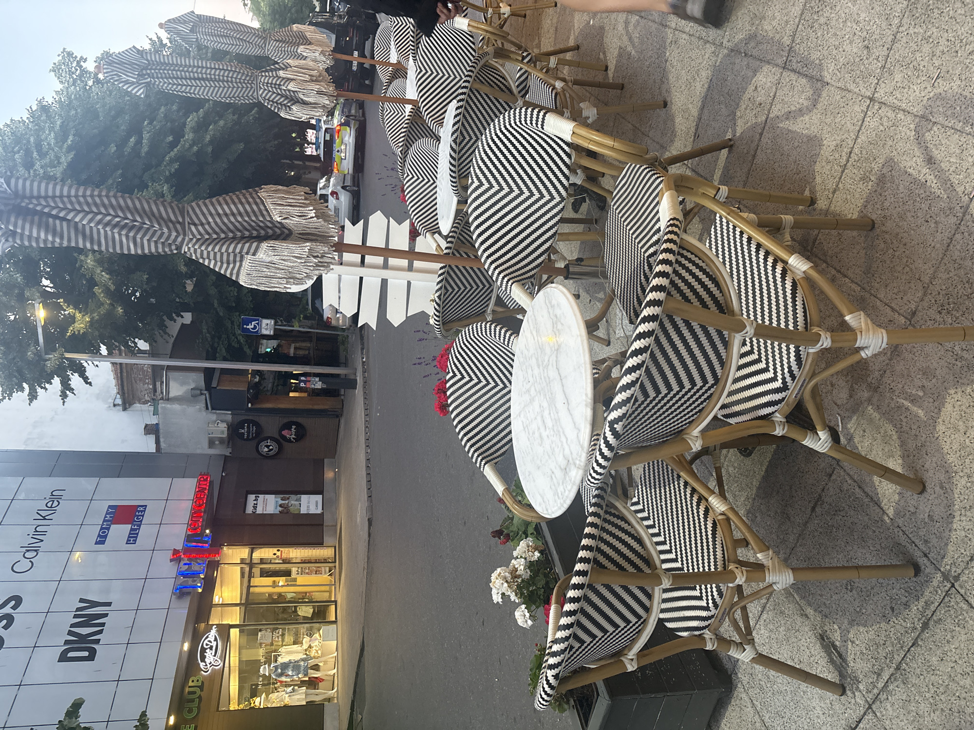 The image captures an outdoor seating area of a cafe or restaurant located on a street in Burgas, Bulgaria. The seating area features several outdoor tables and chairs, all with a distinctive black and white chevron pattern on the seats and backs. The chairs have bamboo-style frames. Small, round marble-top tables are placed between the chairs. In the foreground, there are planters with white and red flowers. Behind the seating area, a street is visible with parked cars, including what appears to be a police vehicle. Several large, striped umbrellas with fringe are set up, some folded and some partially extended. In the background, the facade of a modern building displays various brand logos, including Calvin Klein, DKNY, and Tommy Hilfiger. There are no visible people actively engaged in the scene, though a leg and foot of a person are visible on the right side of the image, suggesting someone is present or has just passed by. The overall scene depicts a commercial street environment in Burgas, likely during daylight hours.